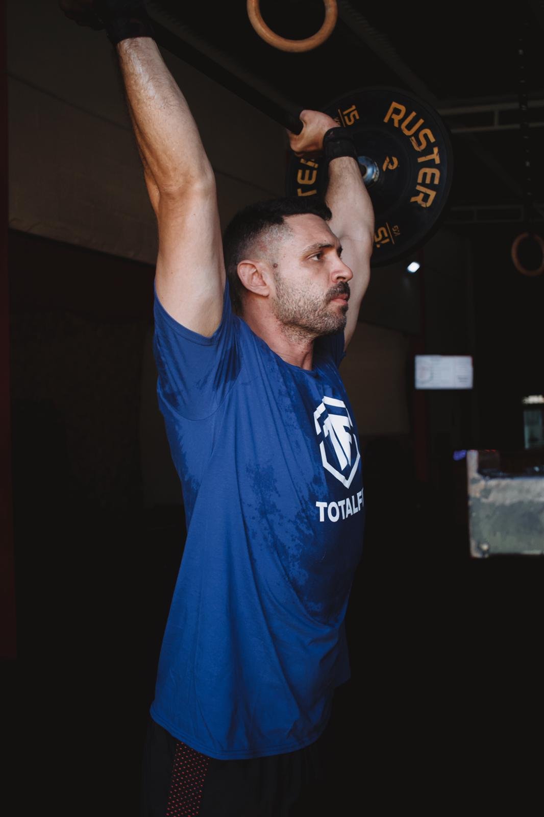 A man in a blue shirt lifting a barbell overhead during a workout in a gym.