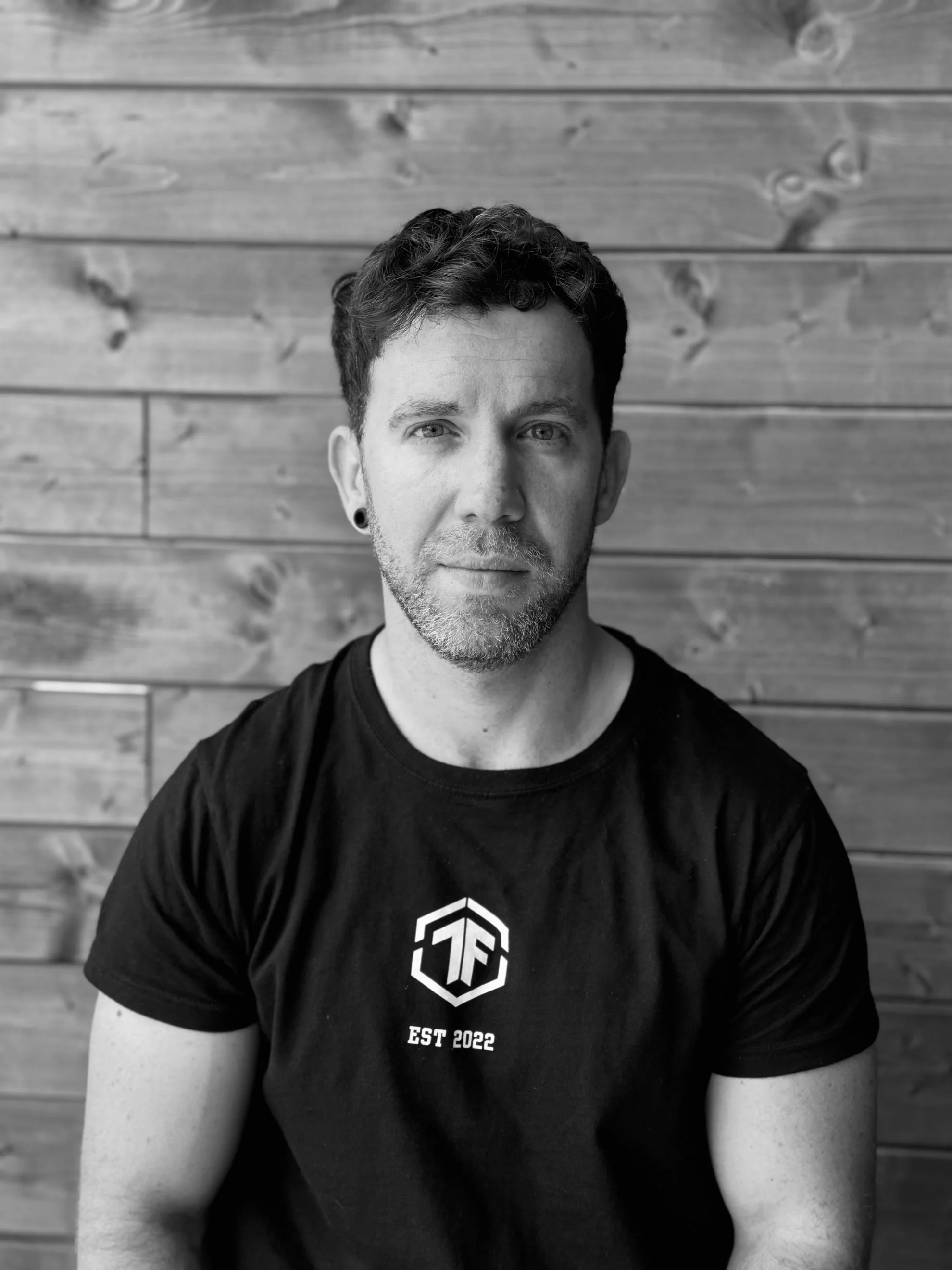 Black and white photo of a man with curly dark hair, beard, and earrings, wearing a T-shirt with a logo and 'EST 2022' text, standing in front of horizontal wooden planks.