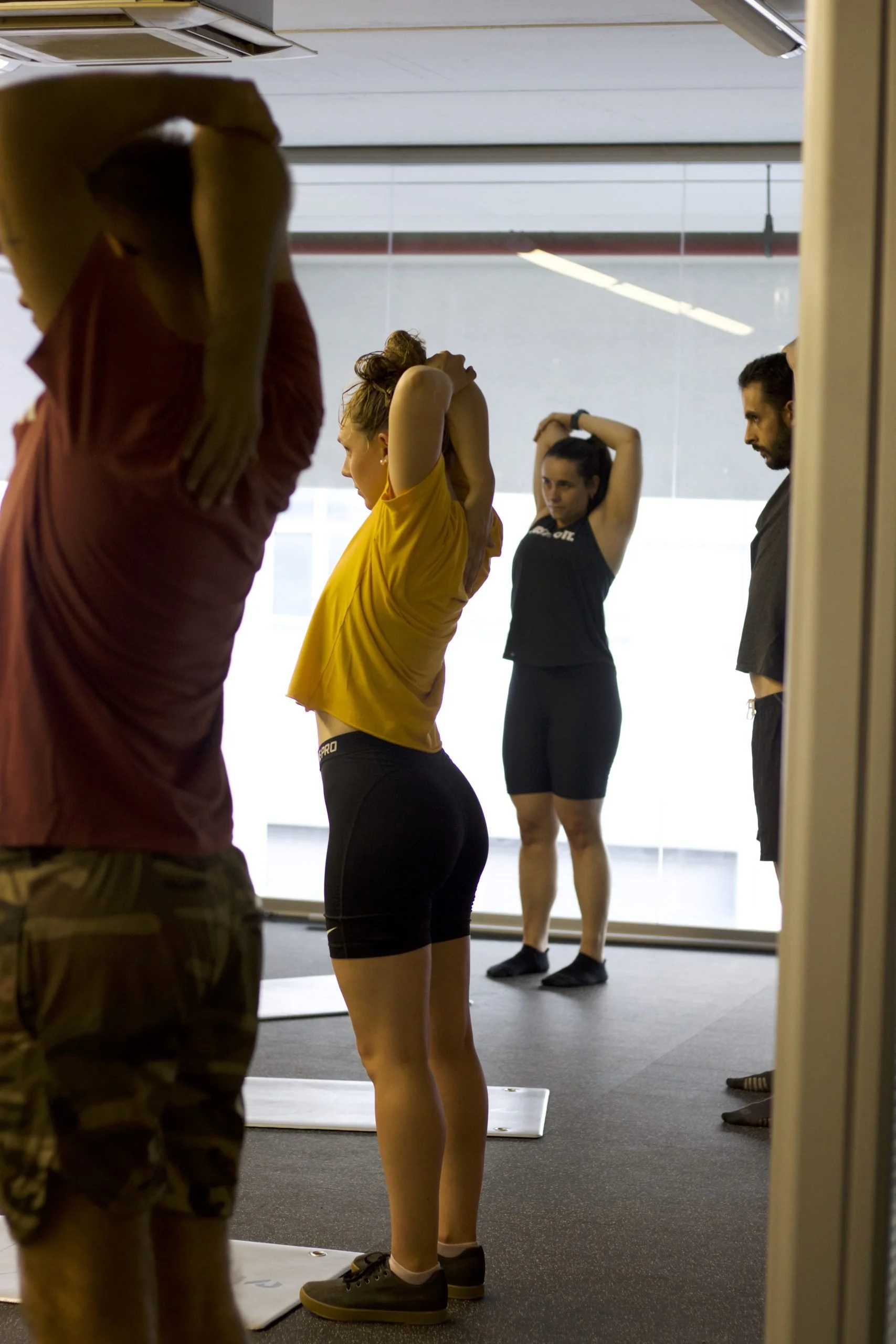 Group of people stretching in a gym, with a woman in a yellow shirt and black shorts in the center, and two men, one on each side, with a woman in a black tank top and black shorts in the background.