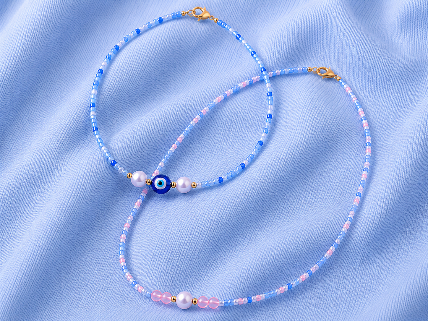 Blue Seed Bead Necklaces with Evil Eye and Pearl Details. Two handmade blue seed bead necklaces on soft blue fabric, featuring pastel pink and white accent beads, gold clasps, pearl details and a blue evil eye centre bead.