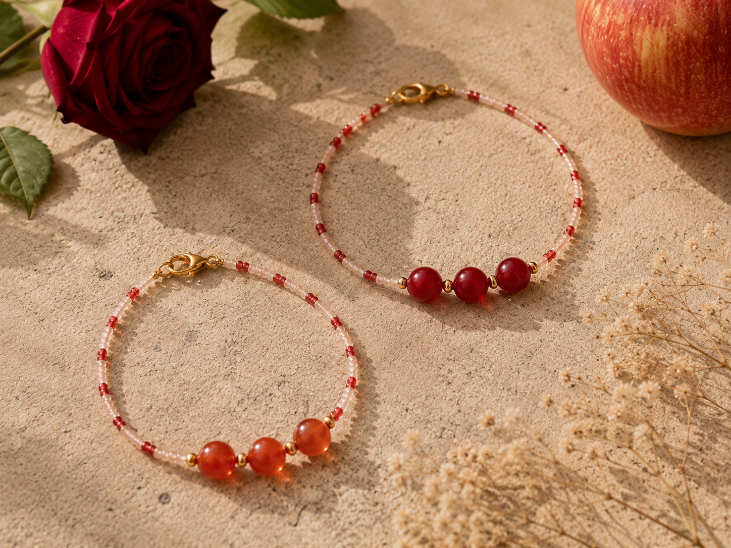 Red Apple-Inspired Beaded Necklaces with Gemstone Details. Two handmade red and blush seed bead necklaces on a warm stone surface, featuring three larger red gemstone beads, gold spacer beads, gold clasps, apple-inspired styling, a red rose and soft 