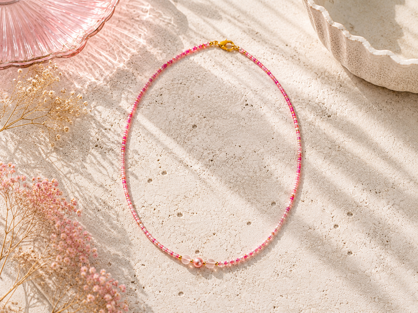 Pink Seed Bead Necklace with Pearl Detail. Handmade pink seed bead necklace on a sunlit stone surface, featuring a pink pearl centre detail, blush accent beads, gold clasp and soft pink floral styling.