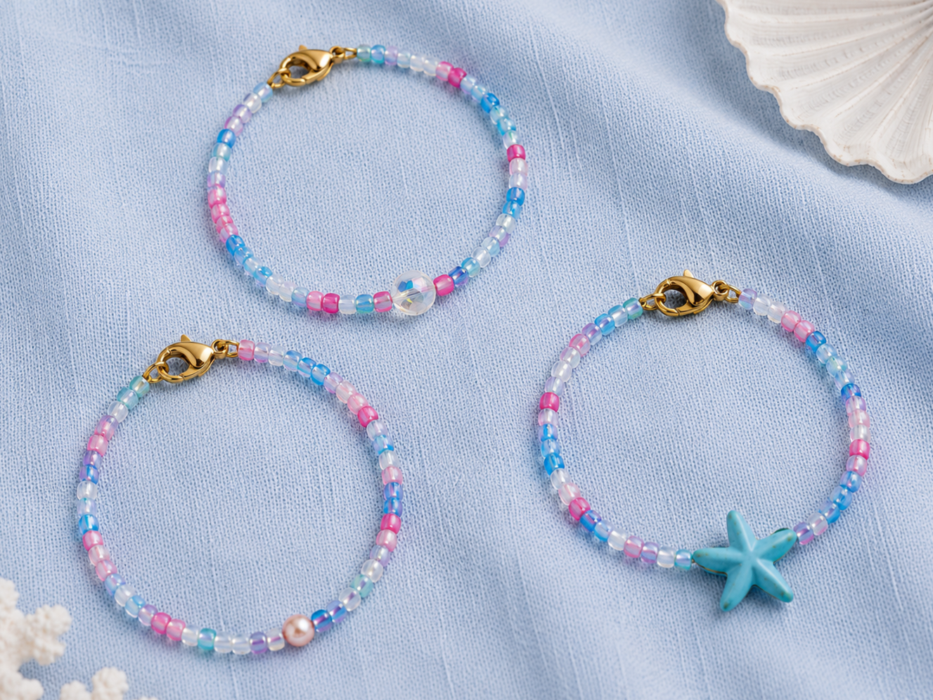 Pastel Mermaid Beaded Bracelets with Starfish Charm | Petals & Pearls. Flat lay of pastel blue, pink and lilac handmade beaded bracelets with iridescent beads and a turquoise starfish charm on soft blue fabric with seashell details.