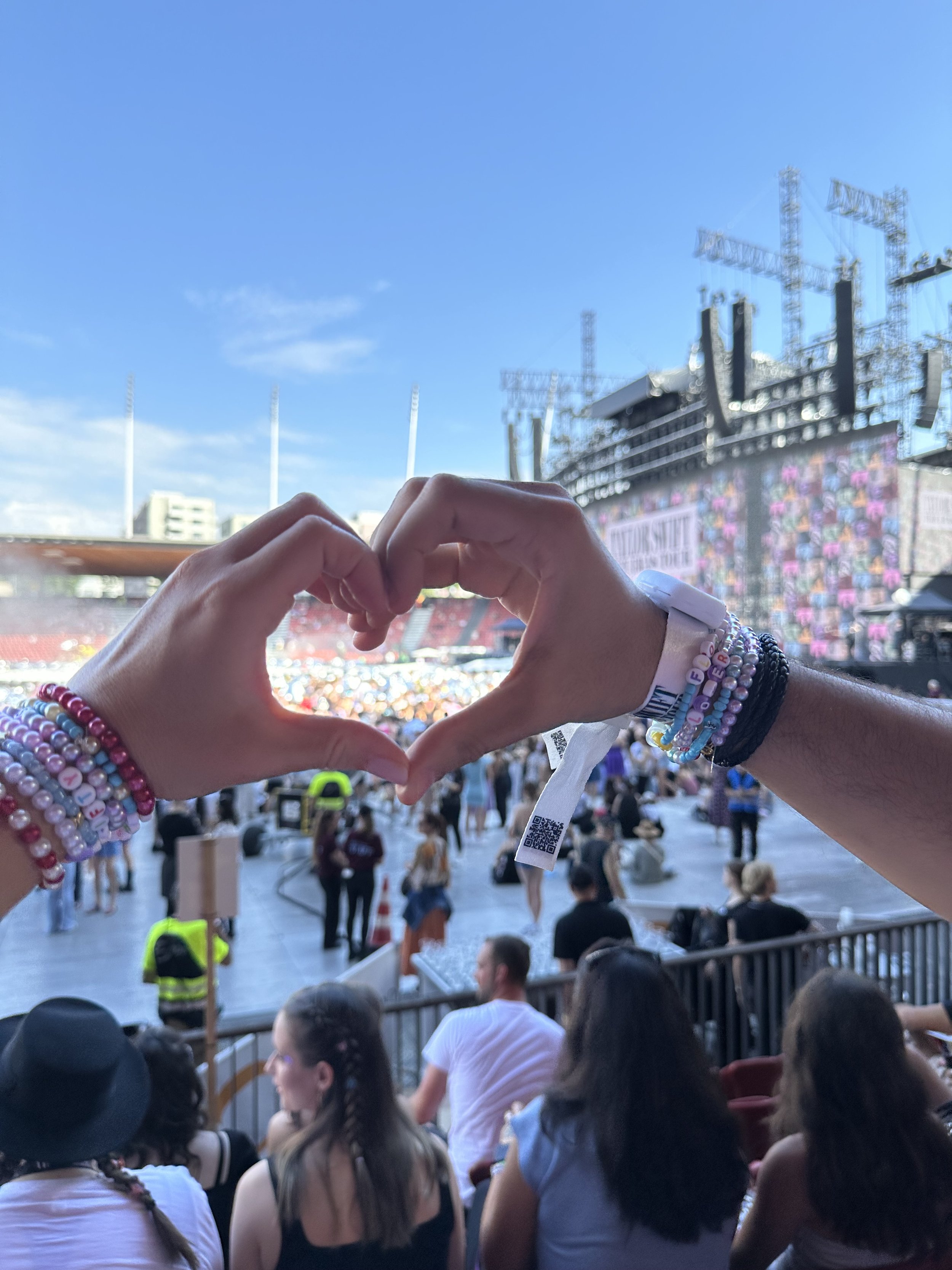Hands making a heart shape at a stadium concert while wearing colourful beaded friendship bracelets, inspired by Petals & Pearls jewellery-making.