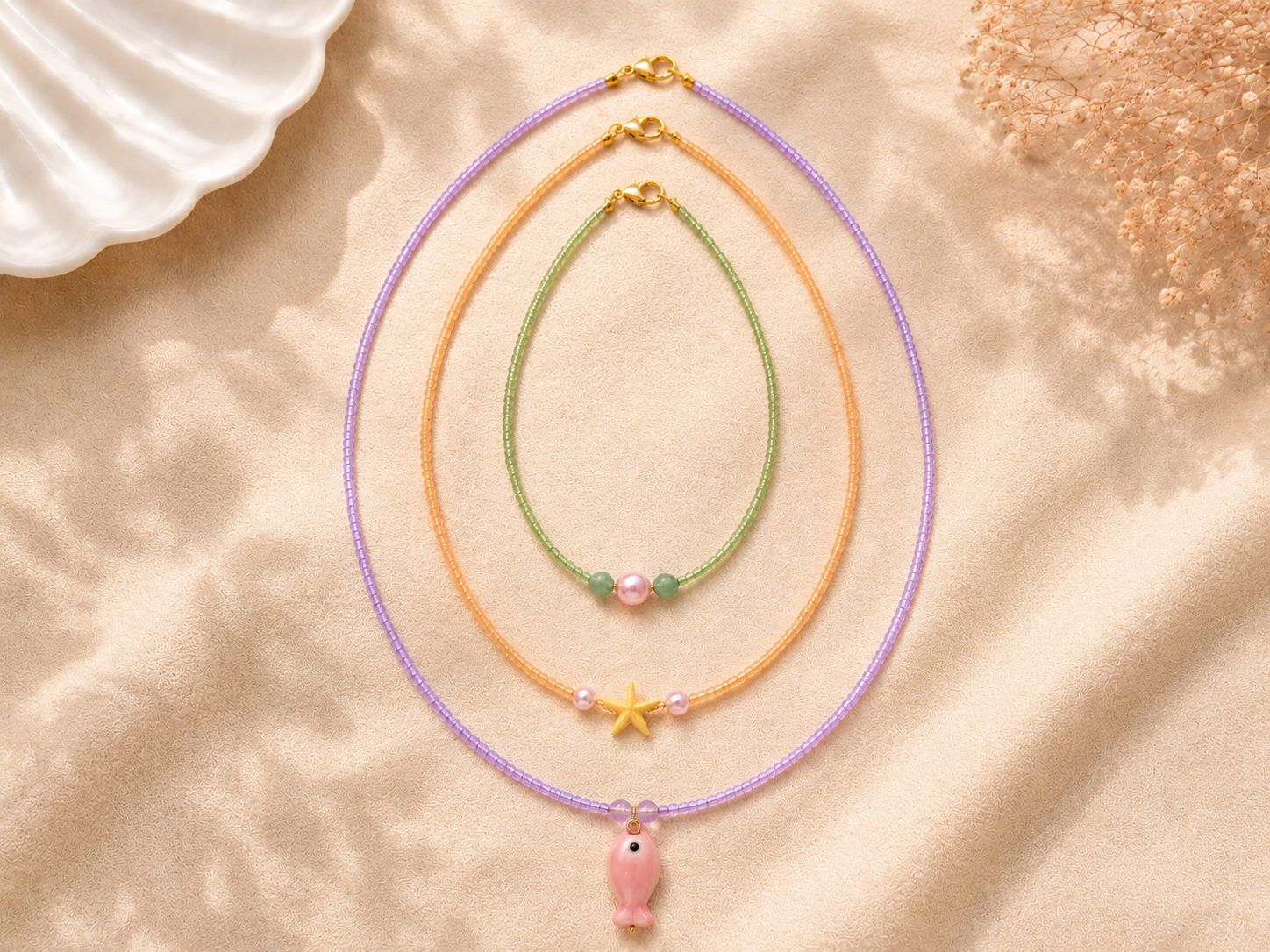 Pastel Layered Seed Bead Necklaces with Starfish and Fish Charms. Three handmade pastel seed bead necklaces on warm cream fabric, featuring lilac, peach and green beads, gold clasps, a yellow starfish charm, pink fish charm, pearl details and soft be