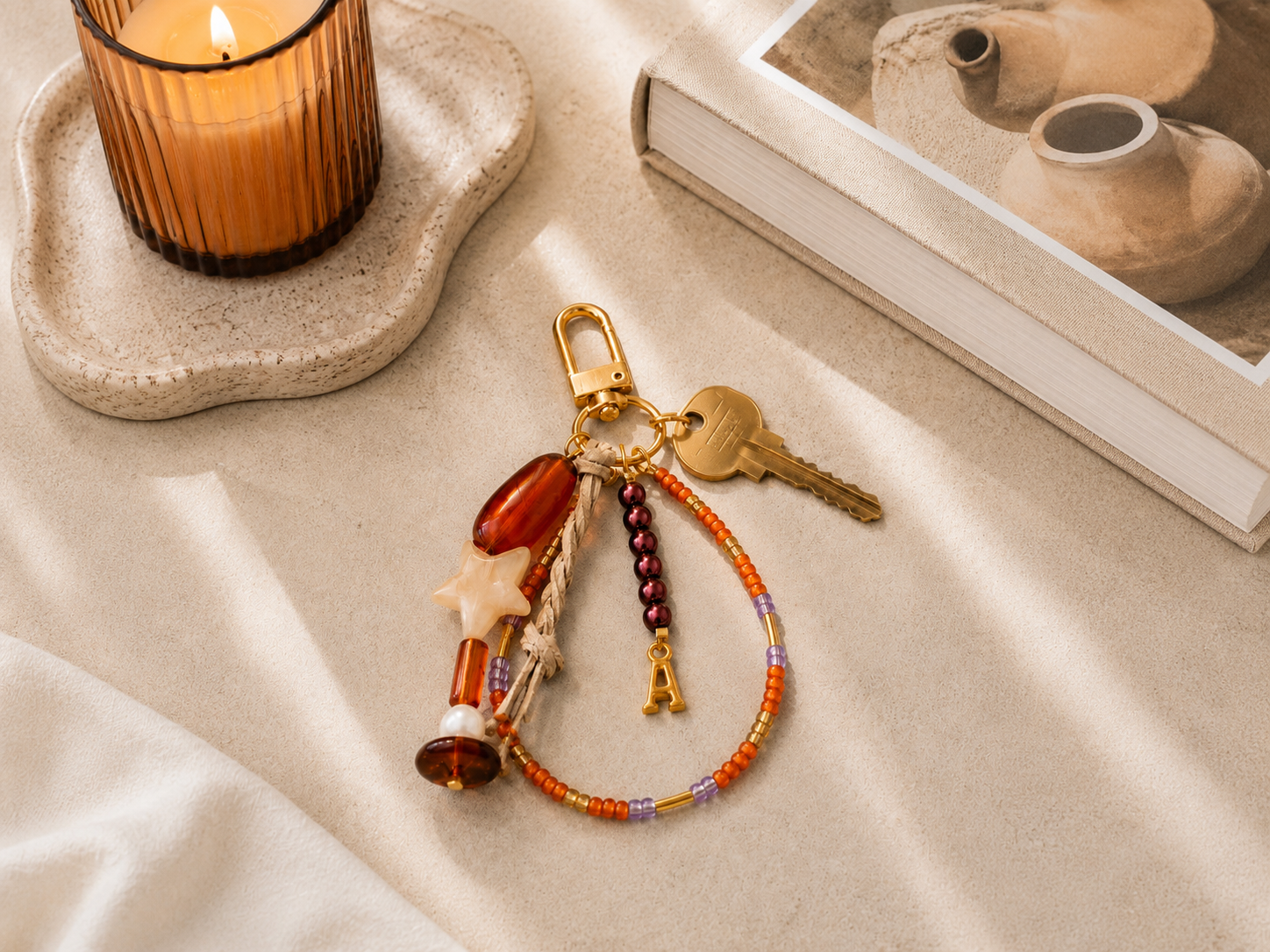 Autumn Beaded House Key Keyring | Petals & Pearls. Autumn-inspired beaded keyring with gold hardware, amber and burgundy beads, cream star charm, gold initial charm and house key styled on a warm neutral tabletop with candlelight.