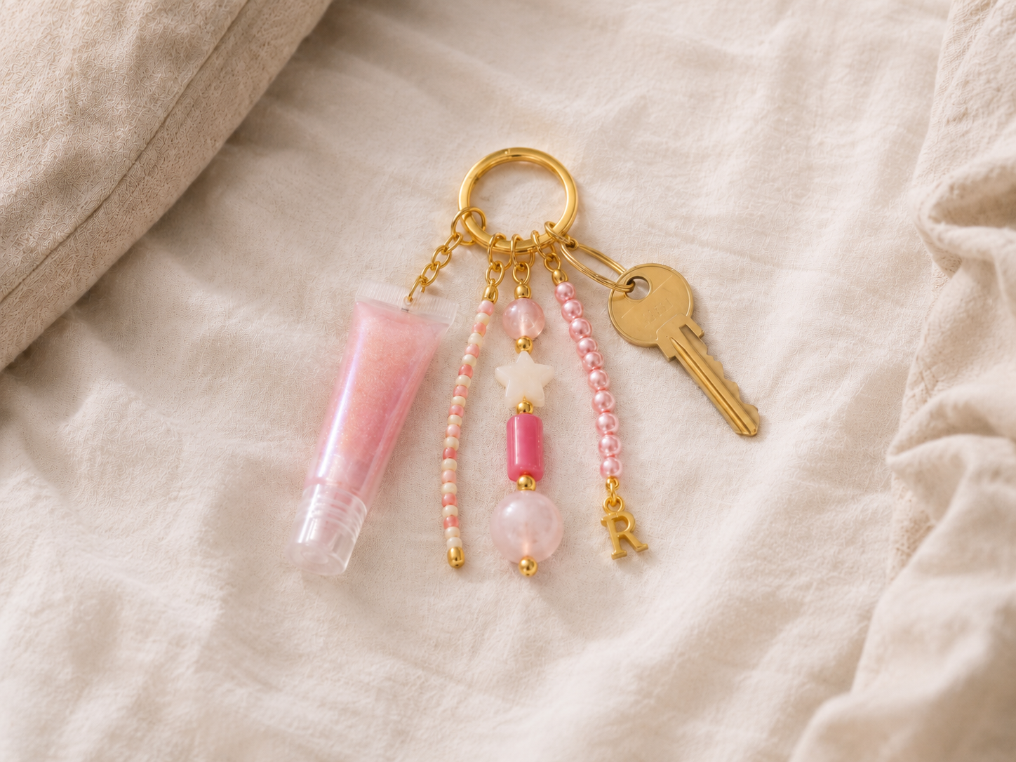 Pink Beaded Keyring with Lip Gloss and Gold Initial Charm. Pink and gold beaded keyring styled on soft cream bedding, featuring a gold keyring, pink lip gloss charm, house key, pearl beads, star bead and gold initial charm.
