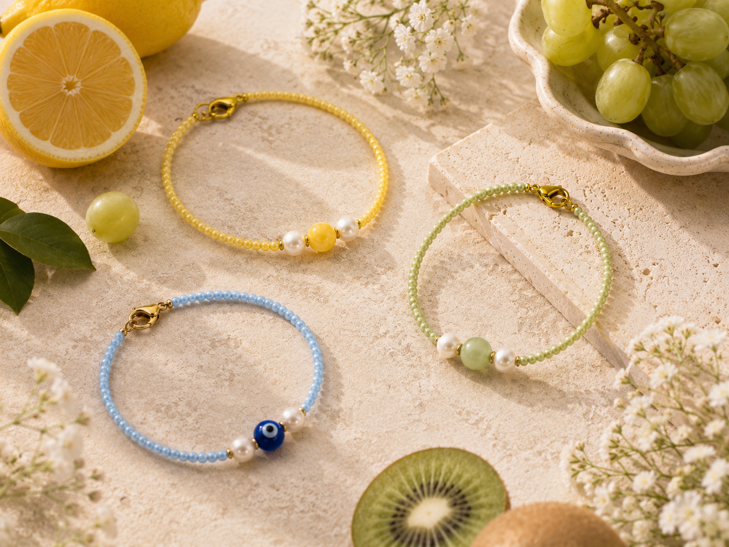 Fruit-Inspired Seed Bead Bracelets with Pearls. Three handmade fruit-inspired seed bead bracelets in yellow, green and blue, styled with pearls, gold clasps, an evil eye bead, lemons, grapes, kiwi and soft white flowers on a sunlit stone surface.