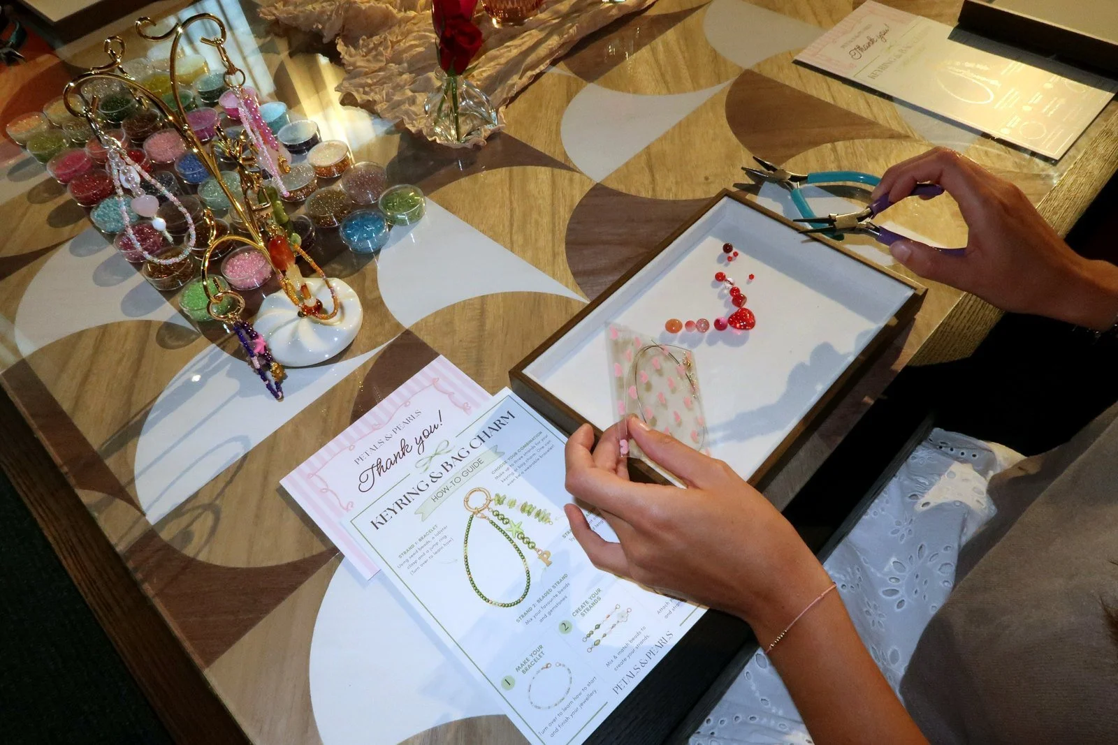 Making a beaded keyring on a design tray at a Petals & Pearls workshop, following the step-by-step guide card.