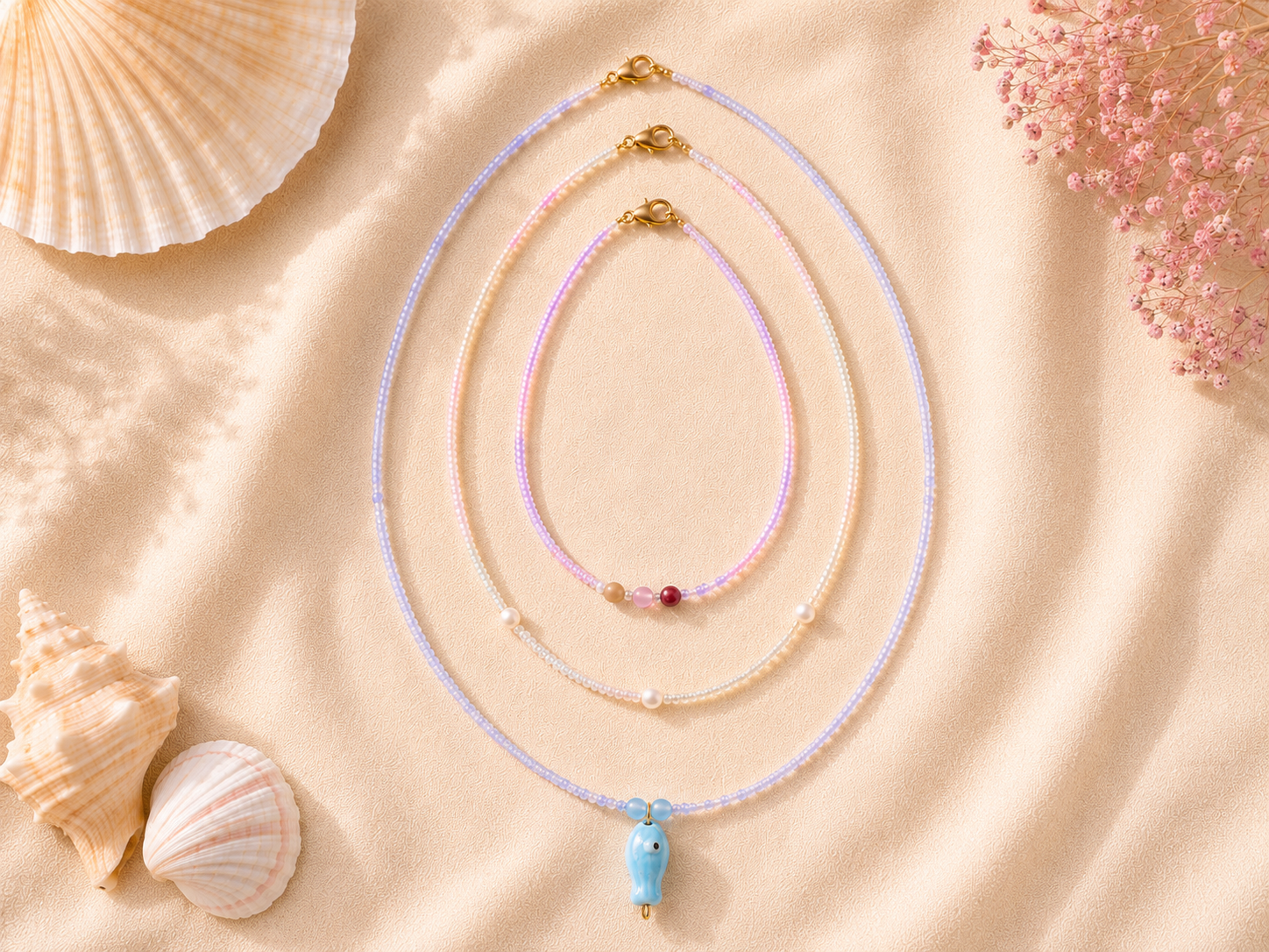 Pastel Layered Seed Bead Necklaces with Blue Fish Charm. Three handmade pastel seed bead necklaces on warm cream fabric, featuring lilac, pink and pearl-toned beads, gold clasps, seashell styling and a blue fish charm.