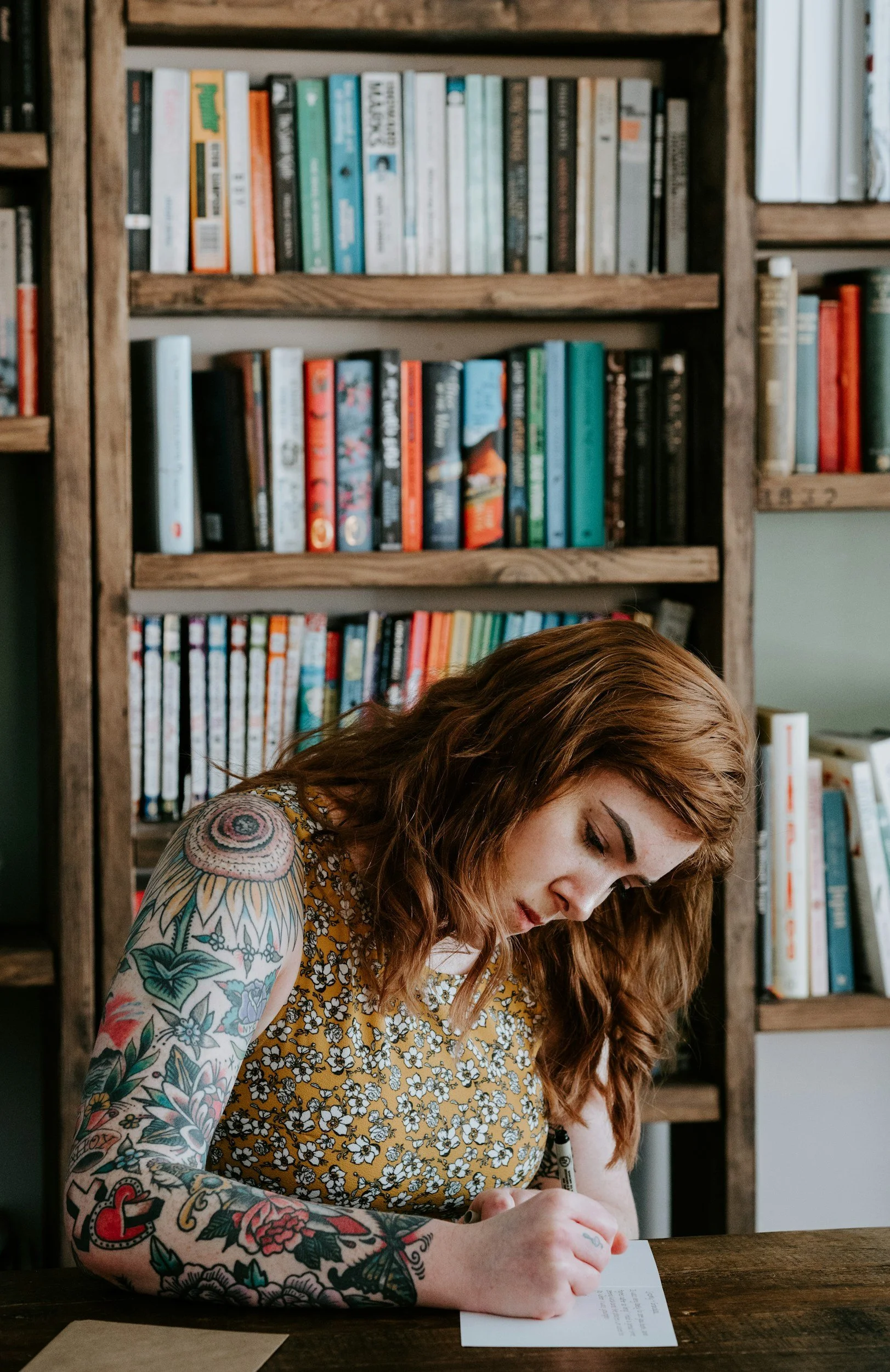 A woman with red hair and colorful tattoos on her arms, wearing a yellow floral dress, sitting at a wooden table, writing on a piece of paper with a pen. Behind her is a wooden bookshelf filled with books.