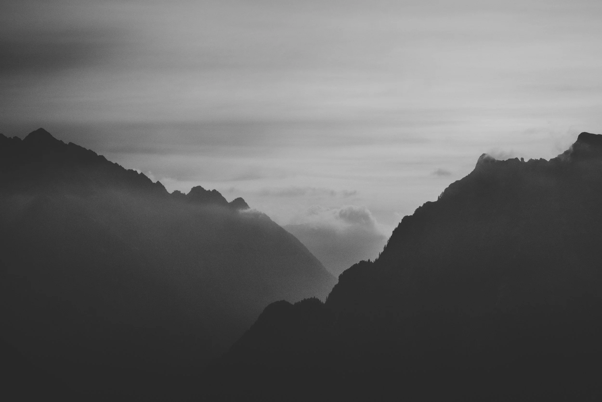 Black and white photograph of mountain ranges with layered silhouettes and clouds, creating a moody and dramatic landscape scene.