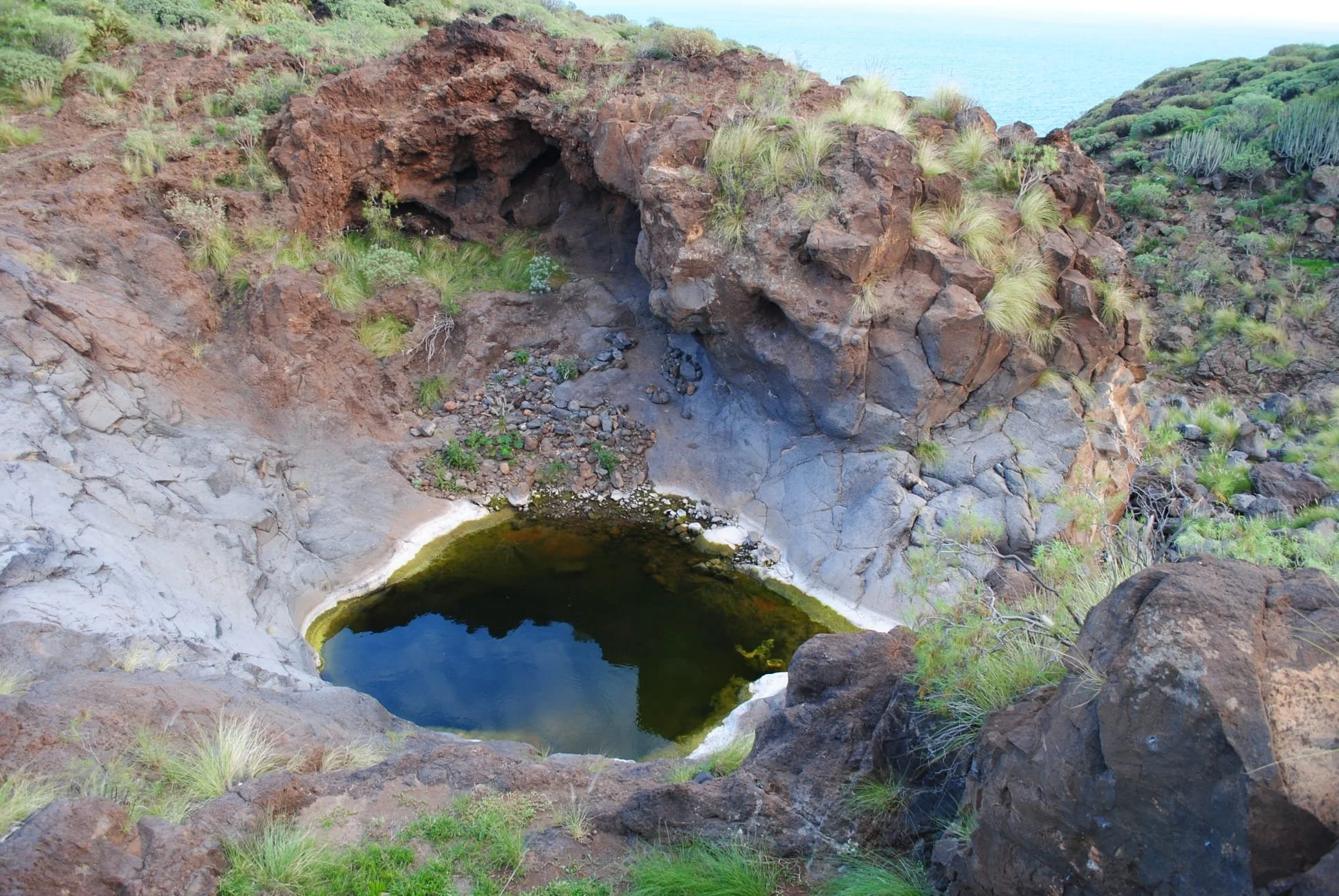 Formación natural en forma de cráter o pozo en el suelo, con agua oscura en su interior, rodeado por rocas y vegetación desertica.