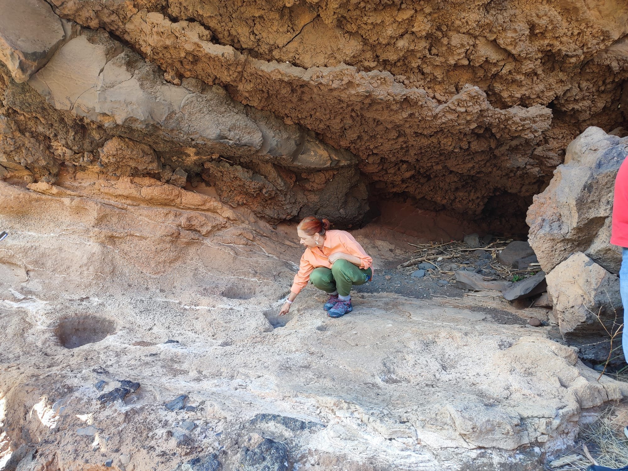 Mujer con camisa naranja y pantalones verdes agachada en un área de excavación, con paredes de tierra y rocas alrededor.
