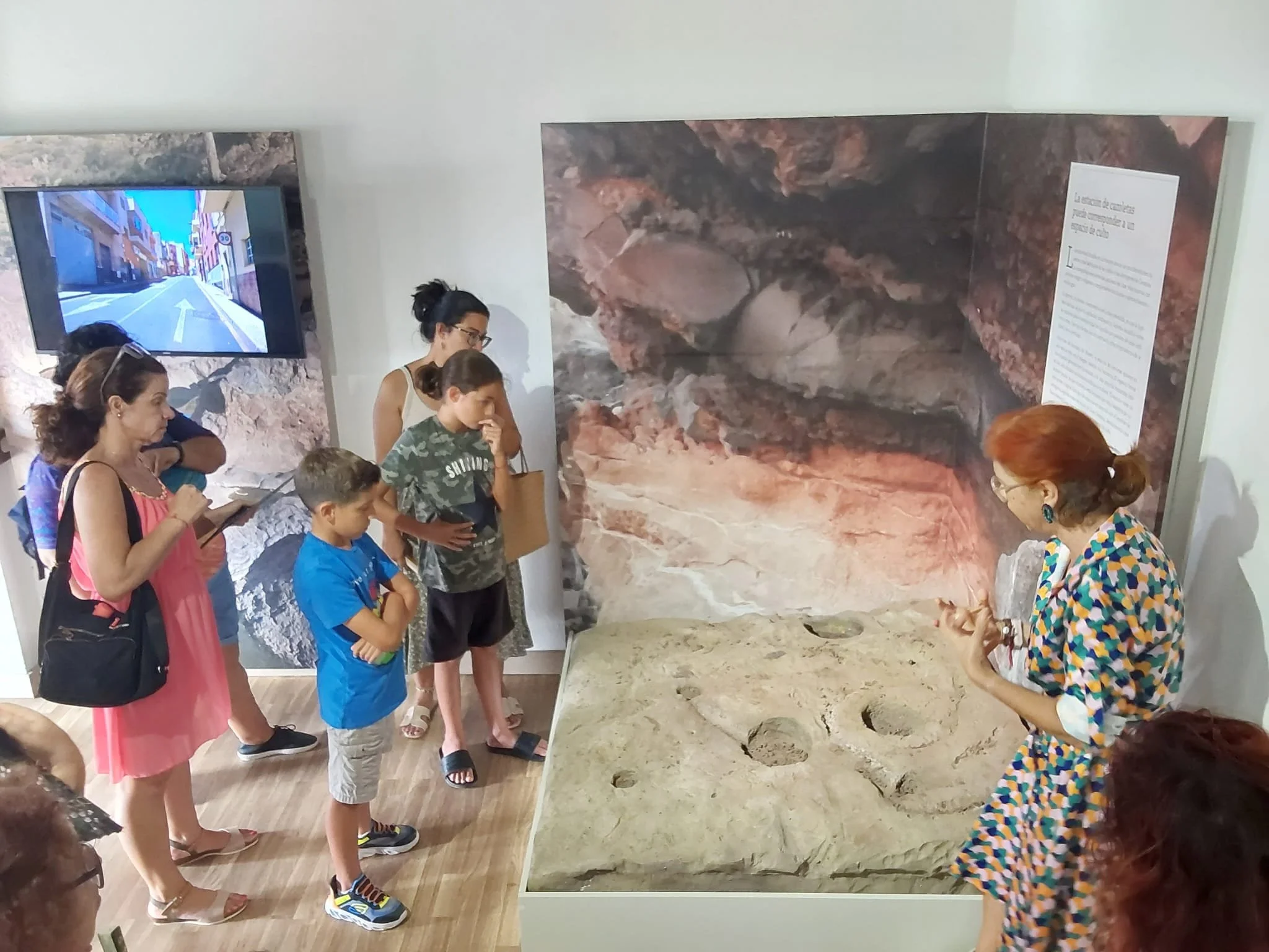 Grupo de personas observando una exhibición en un museo, con un cartel explicativo y una pantalla con imagen de una calle. La exhibición incluye una maqueta en el centro.