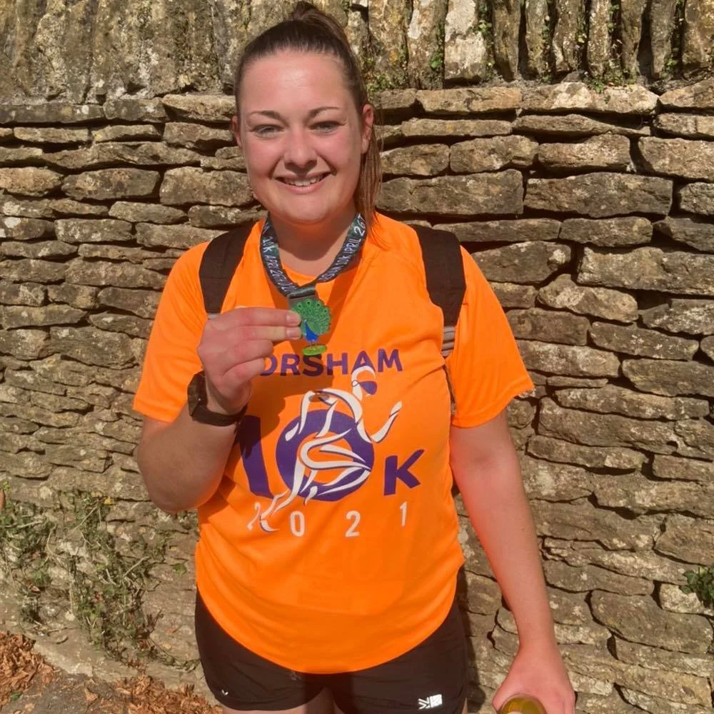 A smiling woman in an orange running shirt holding a medal, standing outdoors in front of a stone wall.
