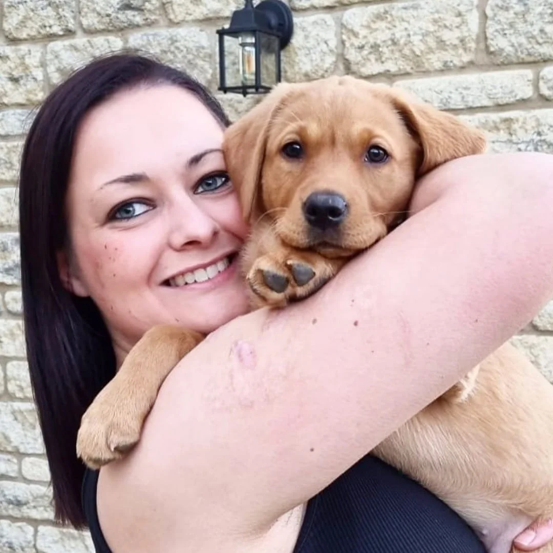 A woman with dark hair and light skin smiling while holding a adorable brown puppy with floppy ears against a brick wall with a black outdoor lantern in the background.