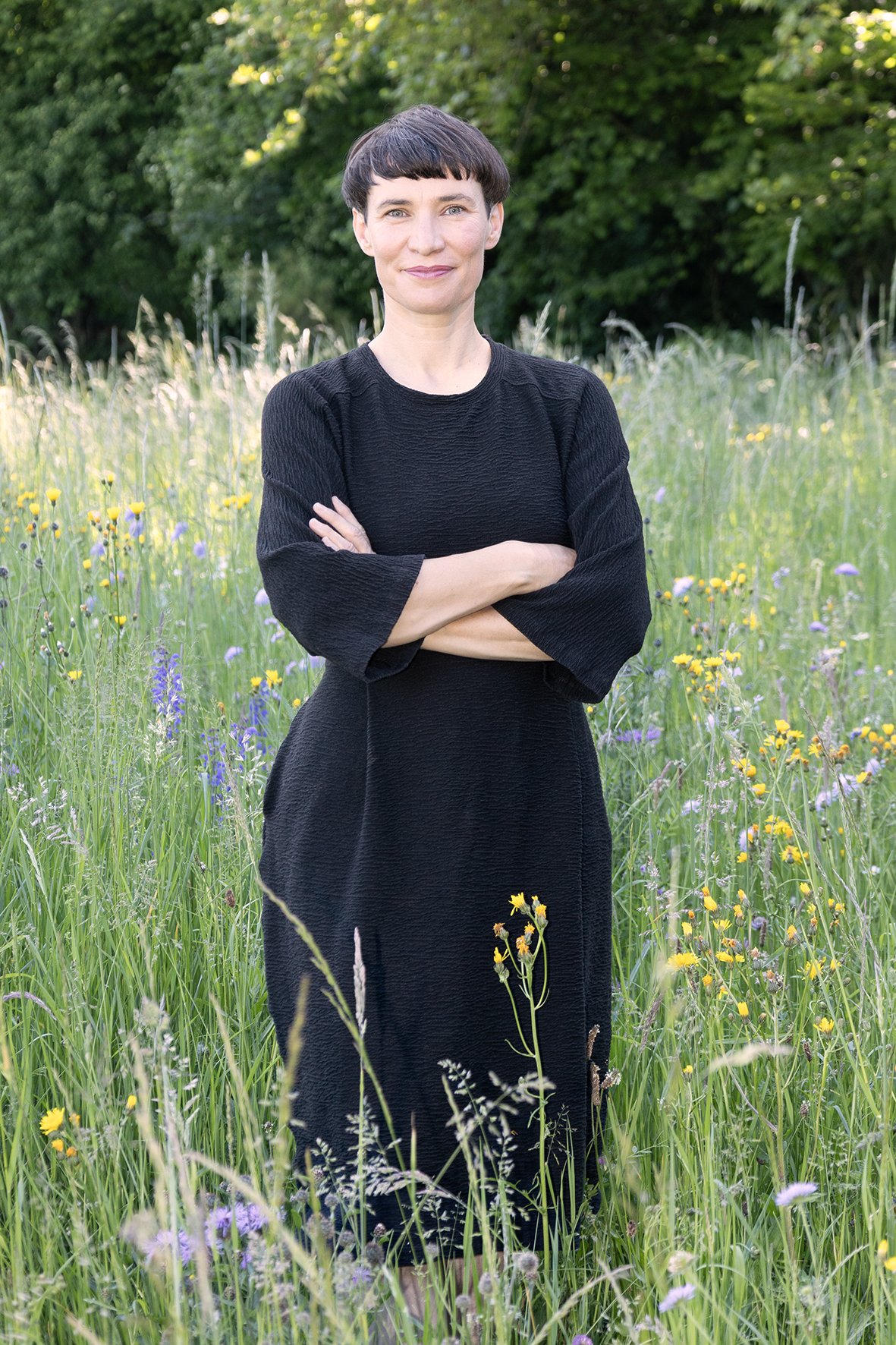 Frau steht mit verschränkten Armen in einem blumenbewachsenen Feld, im Hintergrund Bäume, trägt ein schwarzes Kleid.