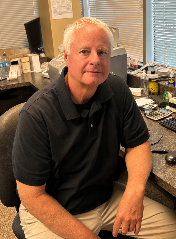 A middle-aged man with light hair, wearing a black polo shirt and beige pants, sitting at a cluttered office desk with various office supplies and equipment, in front of window blinds.