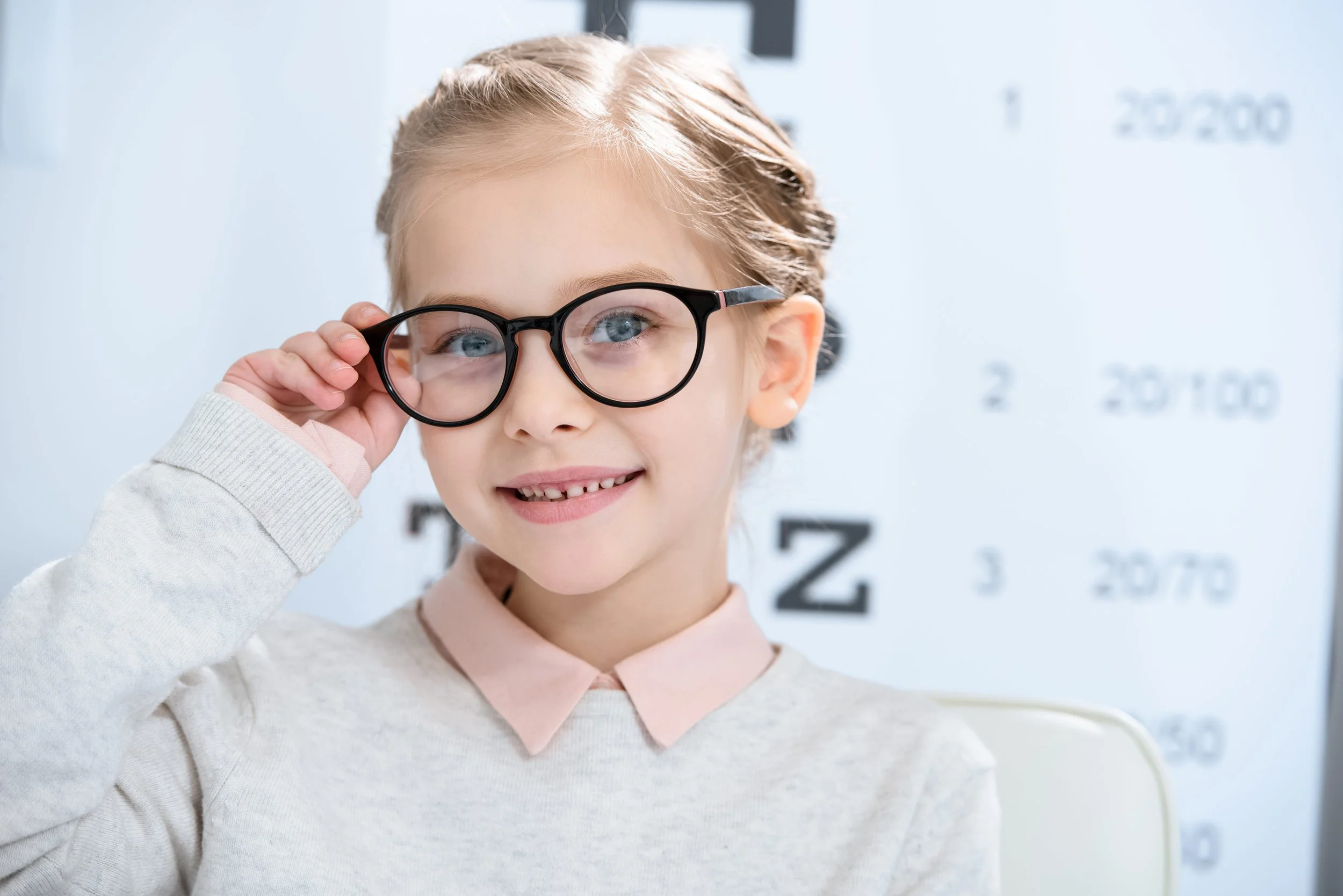 Young girl with blonde hair and blue eyes trying on black glasses, standing in front of an eye chart.