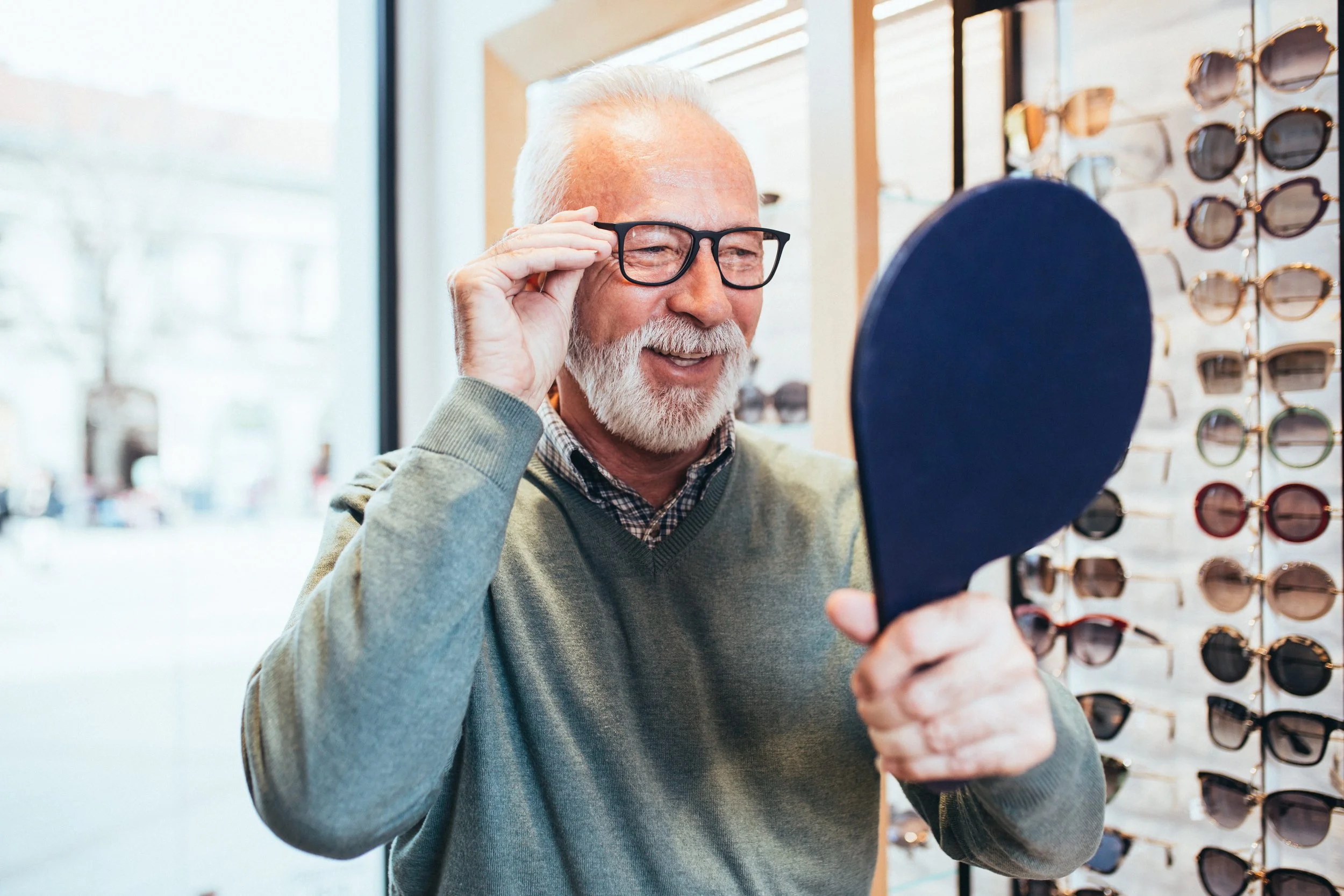 An elderly man with white hair and beard trying on black glasses at an eyewear store, smiling while looking into a mirror.