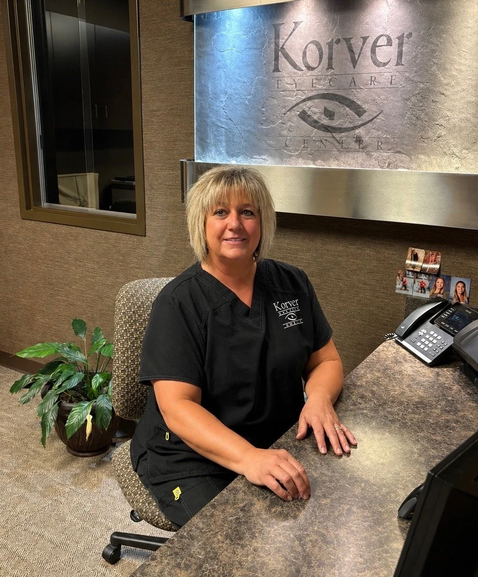 A woman with blonde hair sitting at a reception desk in a medical office, smiling at the camera, wearing a black uniform with the logo 'Korver Eye Care'. Behind her is a sign with the same logo and eye illustration. There is a potted plant on the floor to her left and a telephone on the desk.