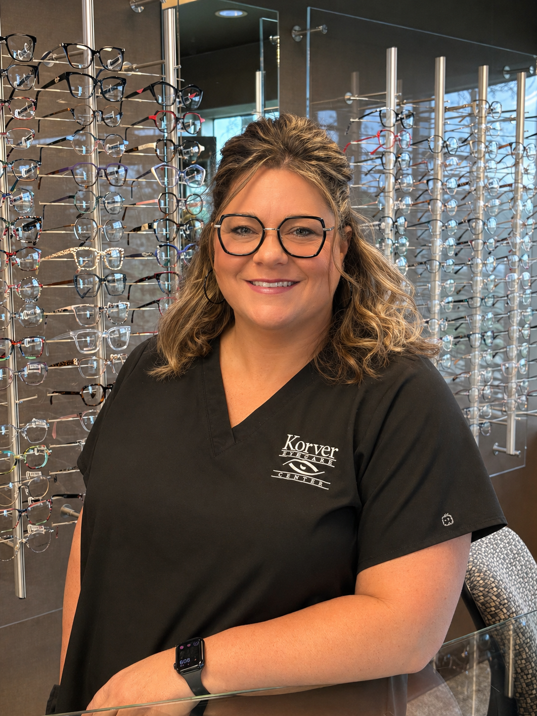 A smiling woman with glasses and wavy hair wearing a black medical scrub top with 'Korver Eye Care Center' logo, standing in front of a display of eyeglasses in an optometry store.