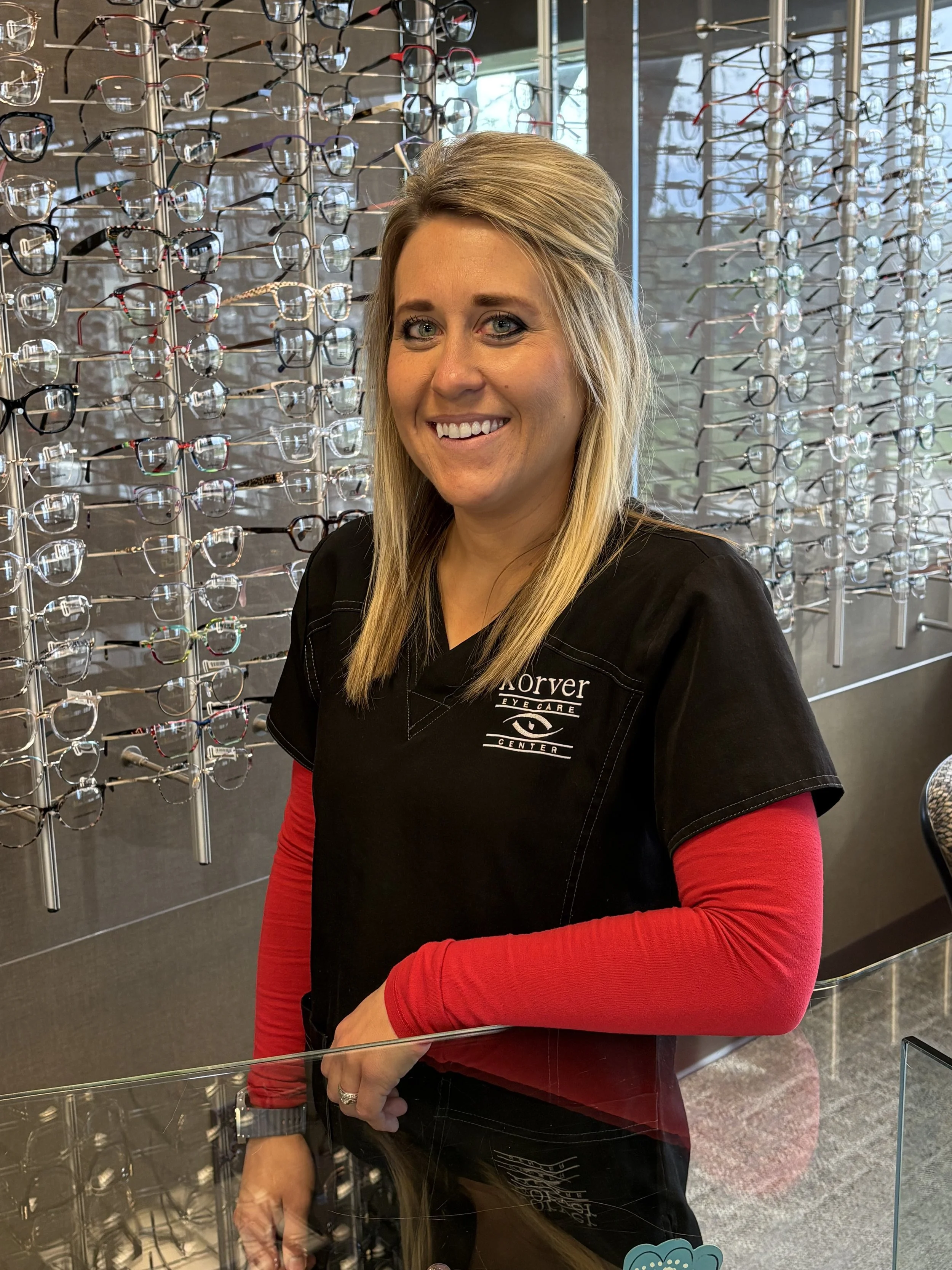 A woman at an eyewear store smiling, standing in front of a display of eyeglasses on a wall.