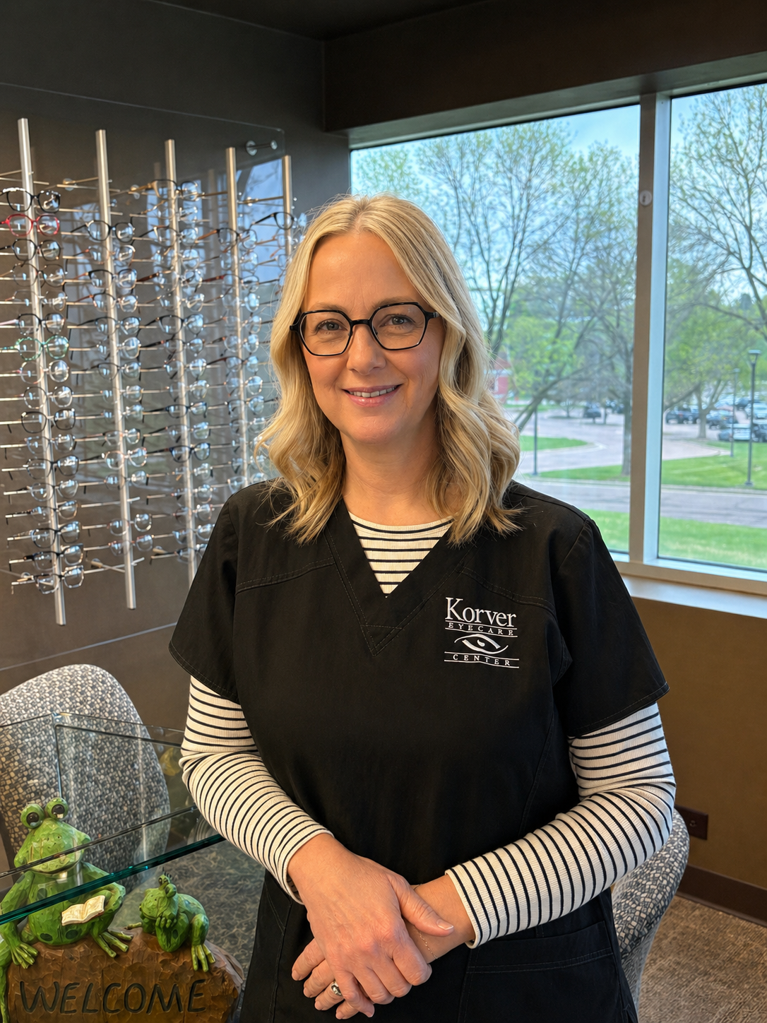 A woman with blonde hair, glasses, and a black uniform with 'Korver Eyecare Center' embroidered on it, standing inside an eye care center, with a display of eyeglasses on the wall behind her and large windows showing trees and a parking lot outside.