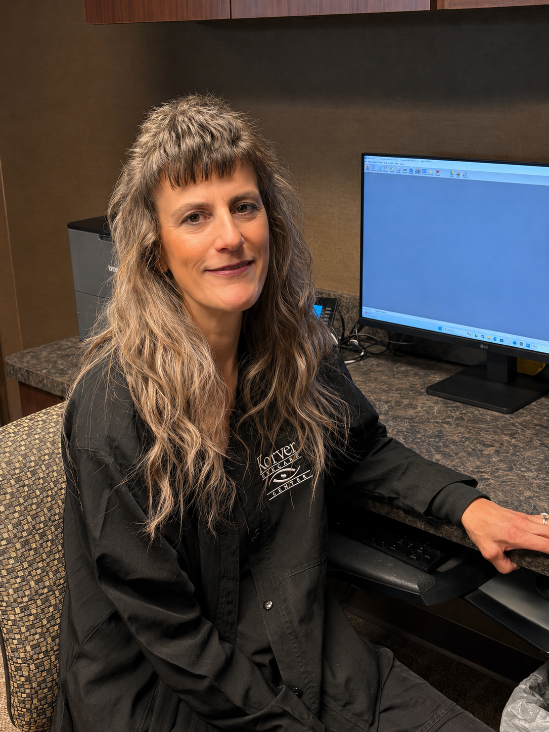 A woman with long wavy hair, wearing a black jacket with white text, is sitting at a desk with a computer monitor in an office setting.