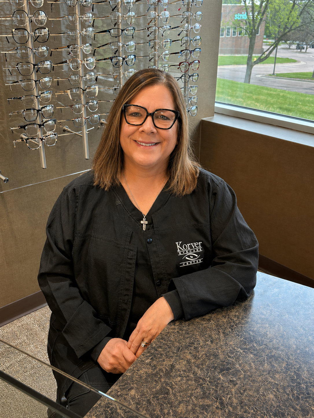 A woman with shoulder-length brown hair wearing glasses and a black jacket with 'Korver Eyecare Center' embroidered on it, sitting at a table inside an optometry store with a display of eyeglasses behind her and a window showing a parking lot and trees outside.