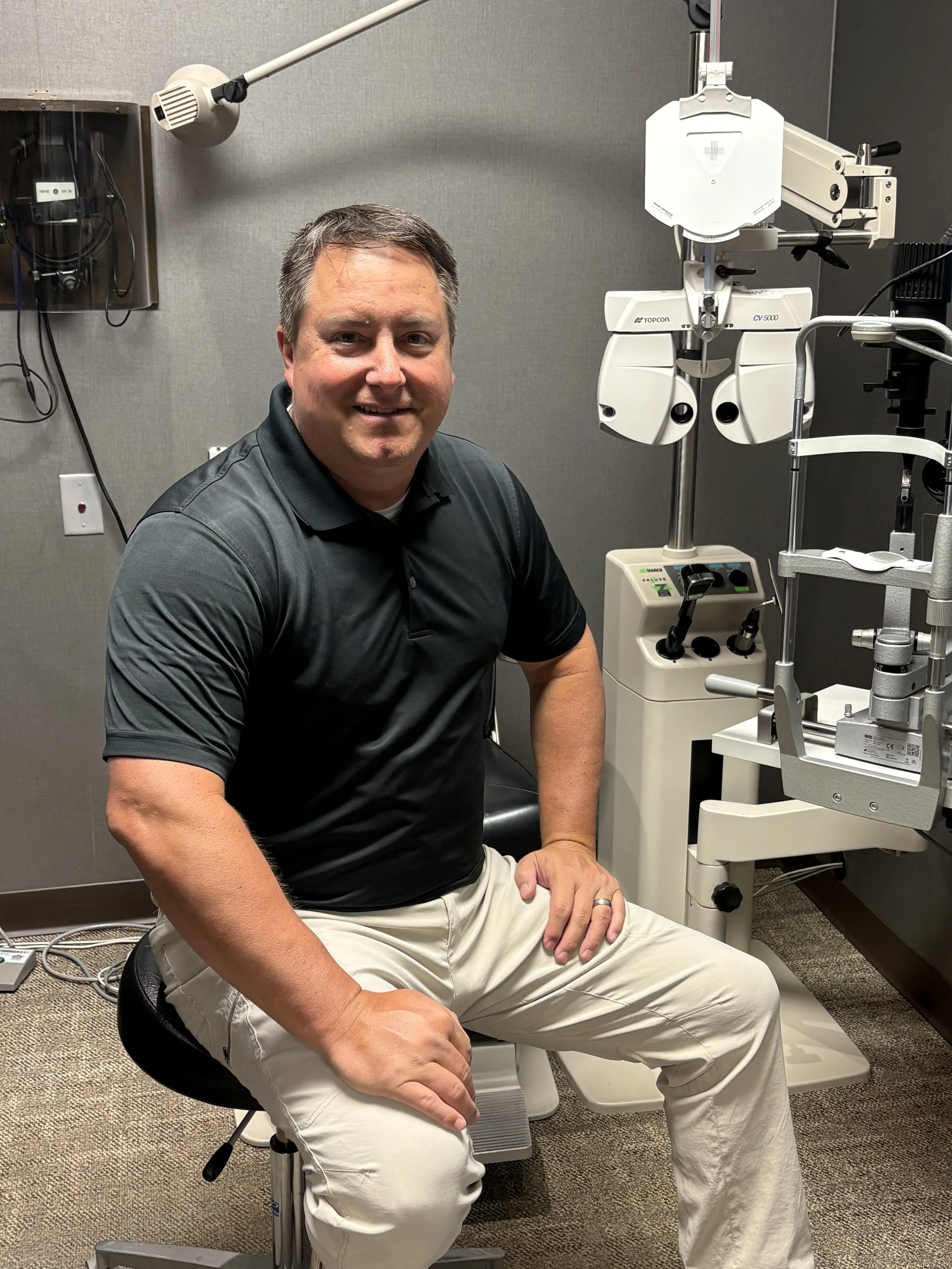 A man sitting in a medical examination room next to ophthalmology equipment, smiling at the camera.