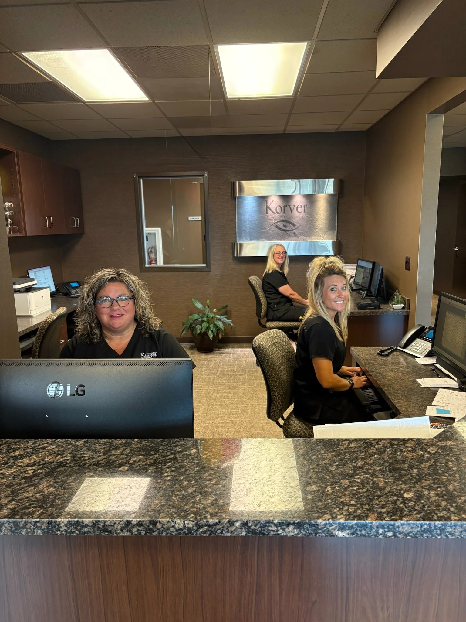 Reception area with three women in black uniforms sitting at desks with computers, a plant, and office supplies, behind a granite counter with a wood panel on the front, in an office or medical building.
