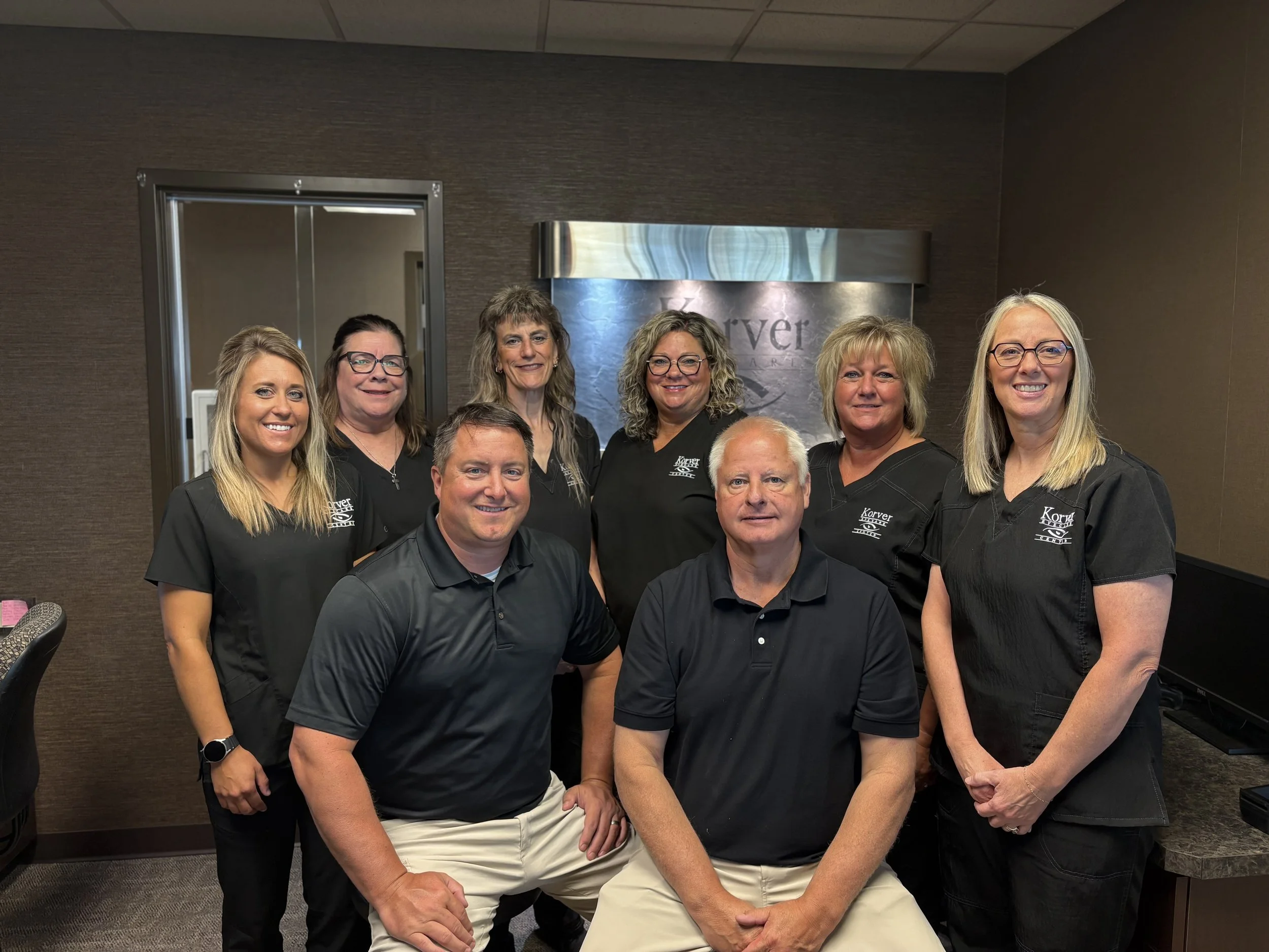 A group of nine people, six women and three men, posing together indoors in front of a wall with a sign that says 'Korrover'. They are all smiling and appear to be dressed in black uniform shirts with white text on the chest.