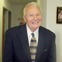 Smiling elderly man in a dark suit and patterned tie, standing indoors near a framed picture.