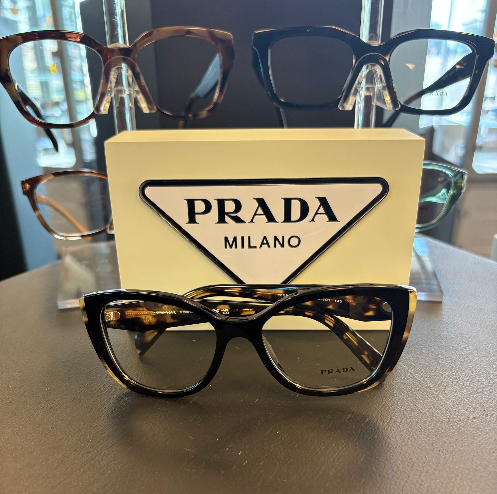 A display of three pairs of designer sunglasses with the Prada Milano logo, arranged on a black surface with a black background and sunlight streaming through windows in the distance.