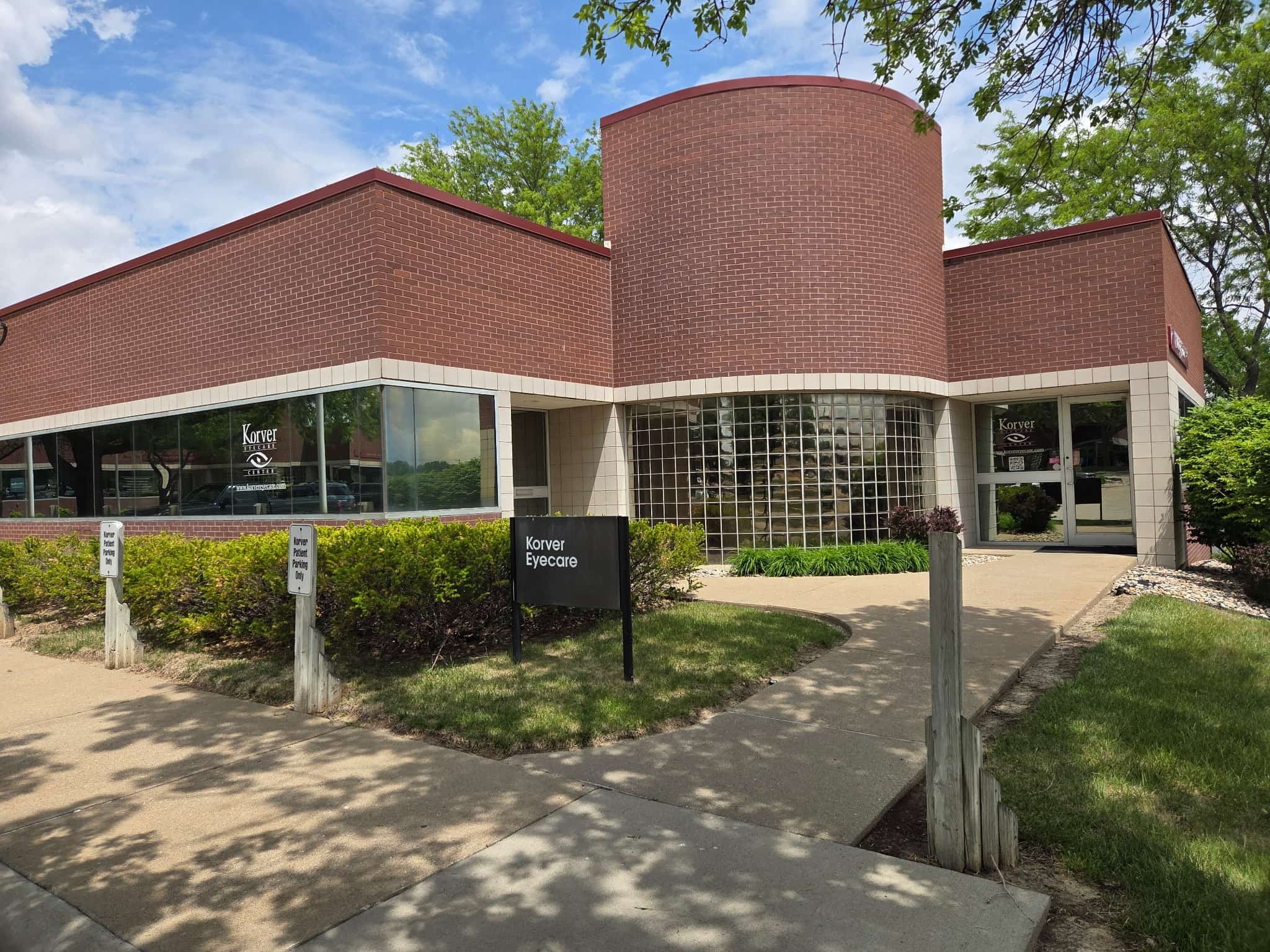 The exterior of a modern building with red brick walls and large glass windows, surrounded by greenery and trees under a partly cloudy sky.