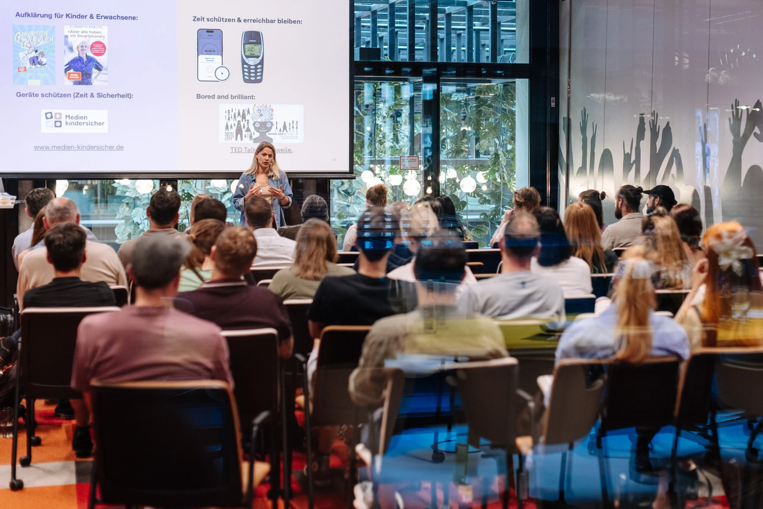Ein Vortrag vor einem Publikum in einem modernen Raum mit großen Fenstern, eine Frau spricht vor einer PowerPoint-Präsentation.