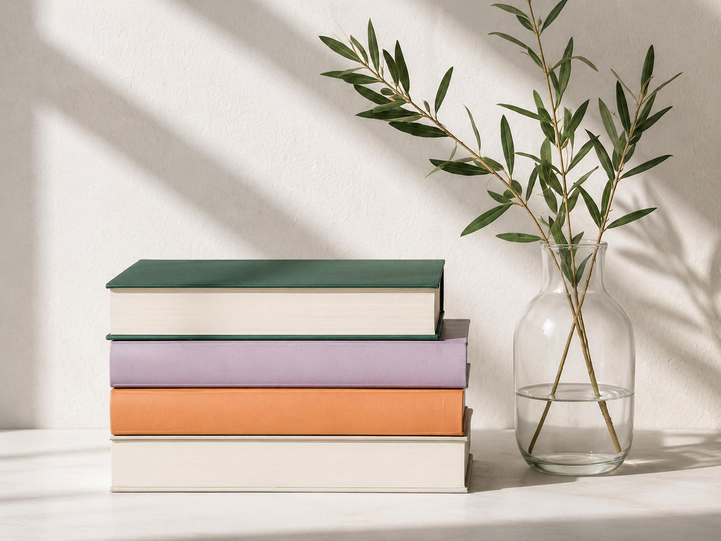 Ein Stapel Bücher mit bunten Einbänden auf einem weißen Tisch neben einer Glasvase mit grünen Zweigen, Sonnenlicht und Schatten an einer weißen Wand.