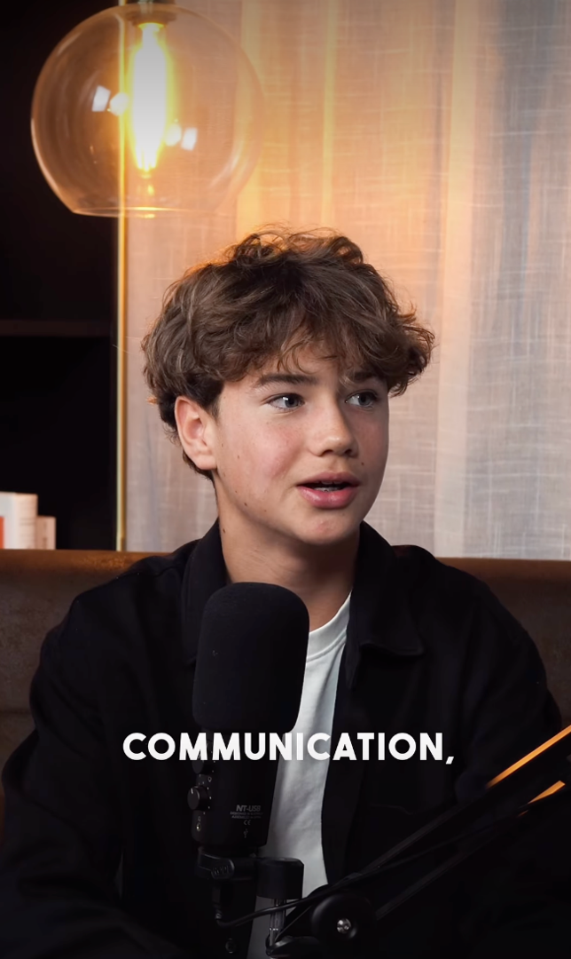 Ein junger Mann spricht in einem Mikrofon während eines Interviews in einem gemütlichen Raum mit warmem Licht und einem Bücherregal im Hintergrund.