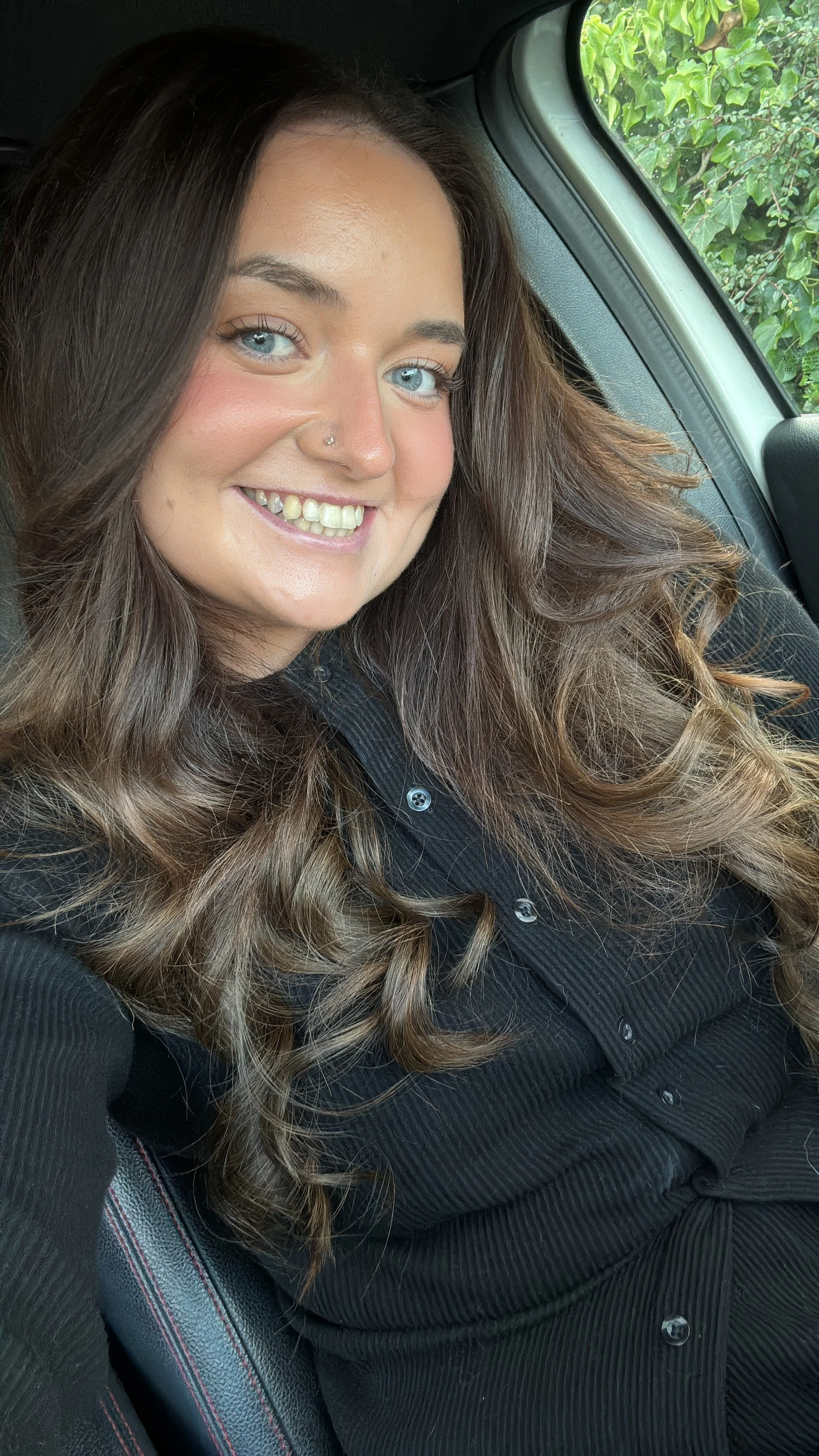 A woman with long, wavy brown hair and blue eyes taking a selfie in a car. She is smiling and wearing a black, button-up shirt with thin vertical stripes. Outside the car window, green leafy trees are visible.