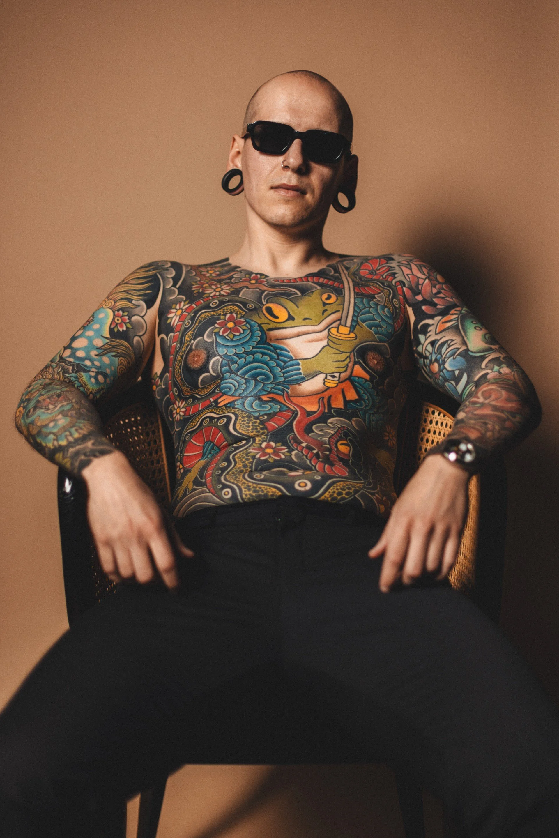 Ein Mensch mit kurzen Haaren, Sonnenbrille, großen Dehnungen im Ohr und vielen bunten Tattoos auf Oberkörper und Armen sitzt entspannt auf einem Stuhl vor einem braunen Hintergrund.