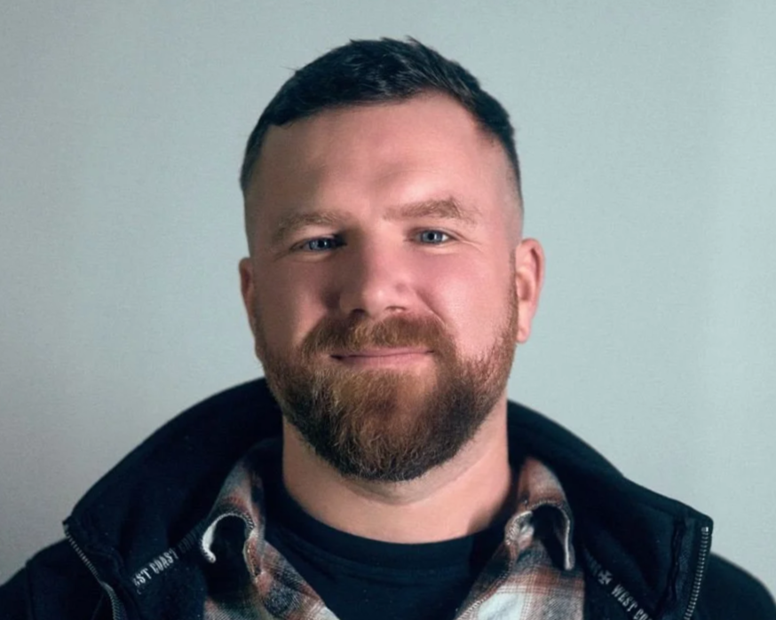 Ein Mann mit dunklen Haaren, Bart und blauen Augen trägt eine schwarze Jacke und ein kariertes Hemd. Er steht vor einer hellen Wand.