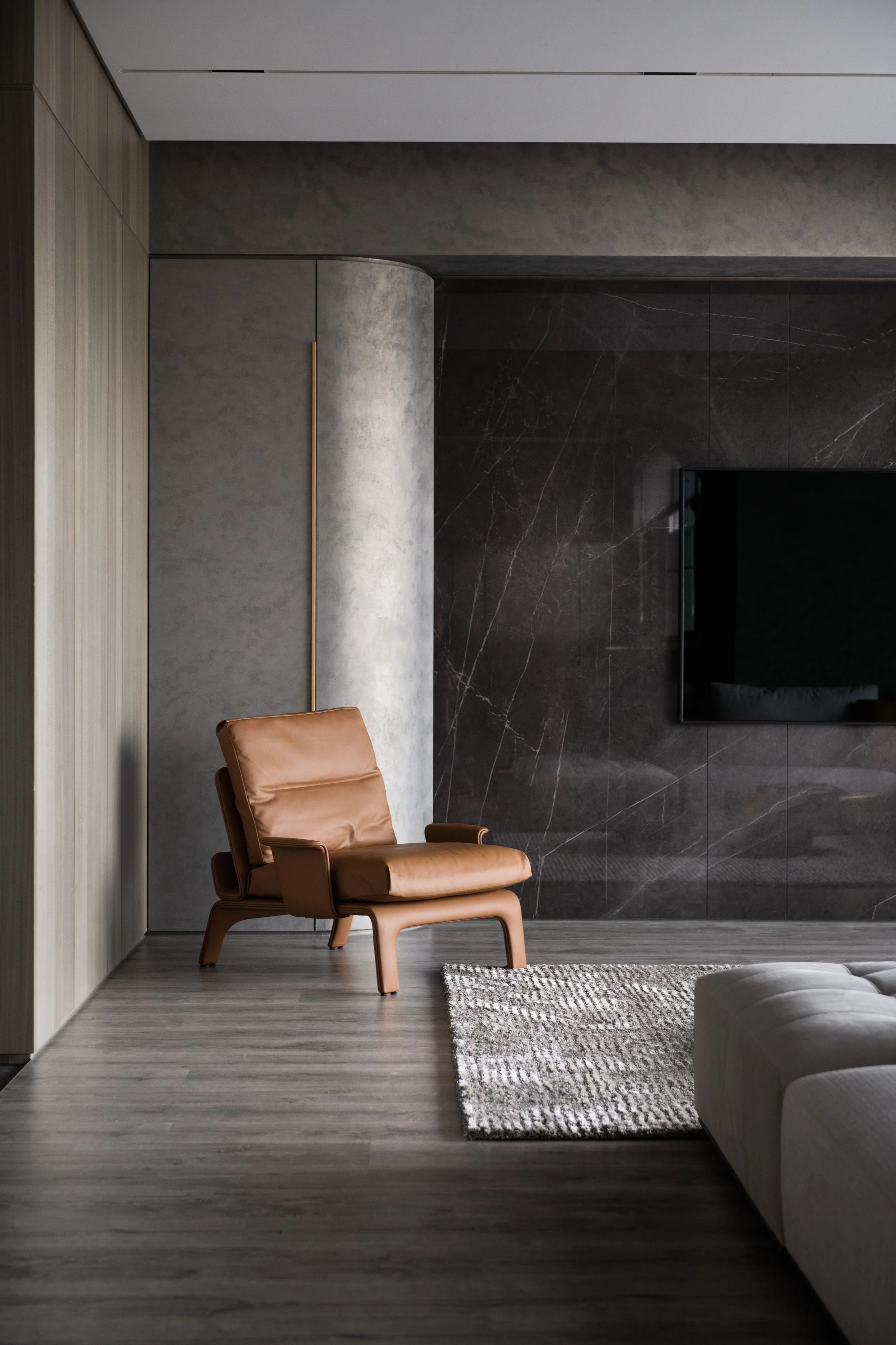 Contemporary living room with a tan leather chair, wooden flooring, a patterned rug, and a large mounted flat-screen TV on dark marble wall.