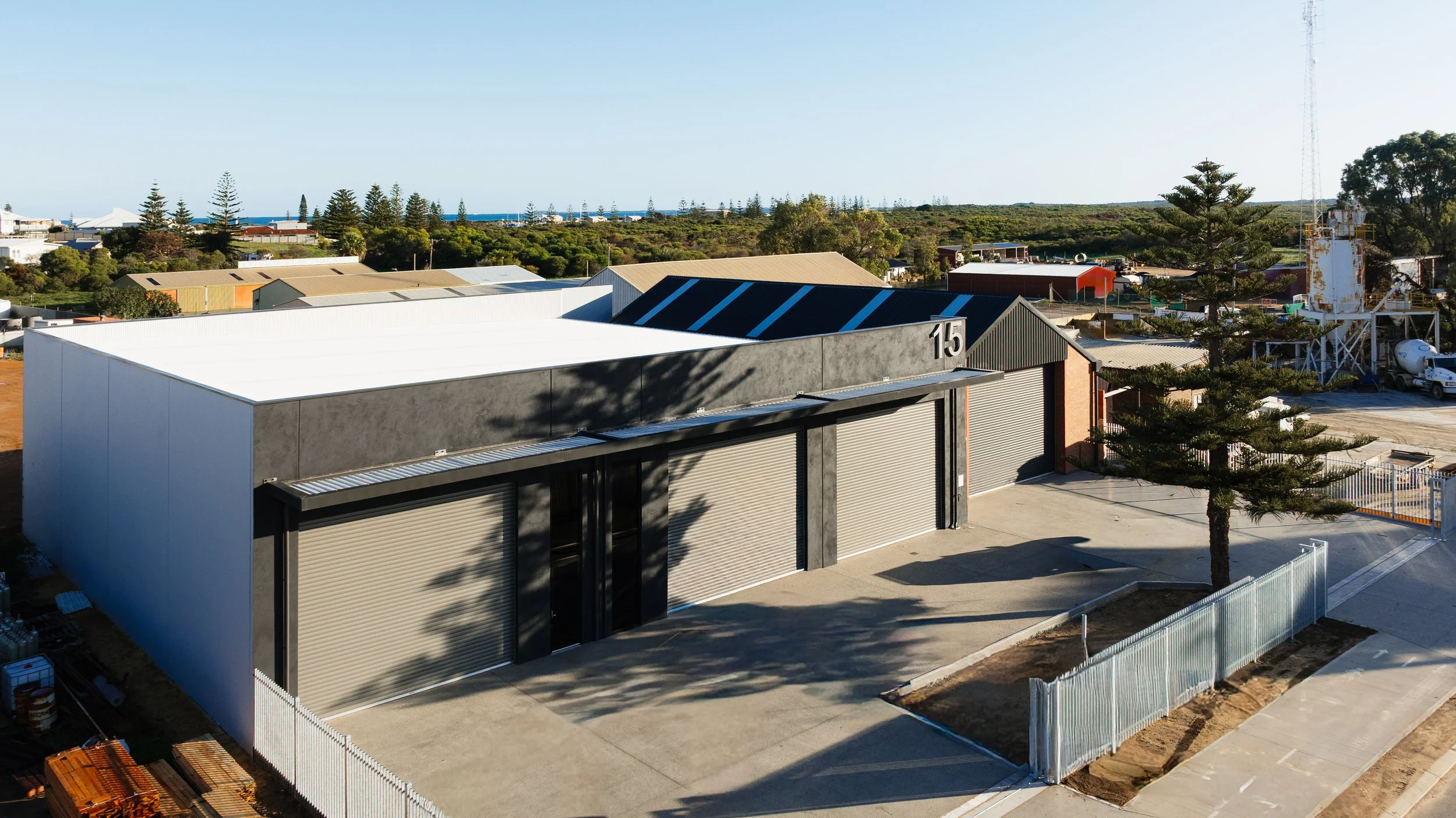Industrial warehouse with metal roller doors in an industrial area near residential homes and lush trees, with a clear sky and some distant water.