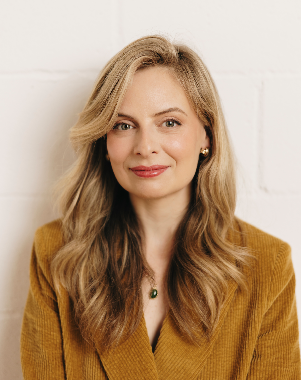 A woman with long wavy light brown hair, wearing a mustard yellow jacket, gold earrings, and a green pendant necklace, smiling at the camera in front of a white brick wall.