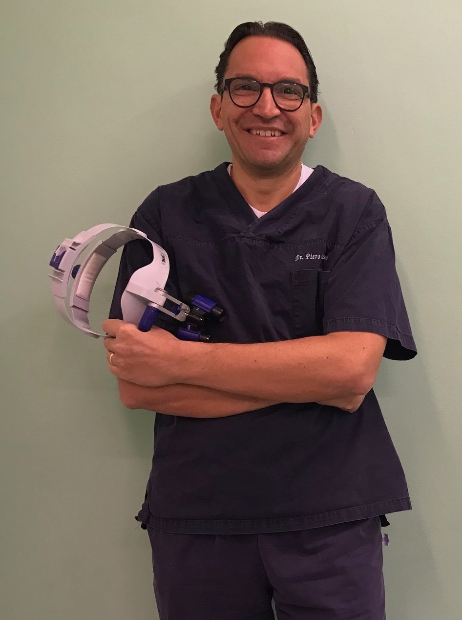 A man in scrubs holding a headlamp, standing against a light green wall, smiling at the camera.