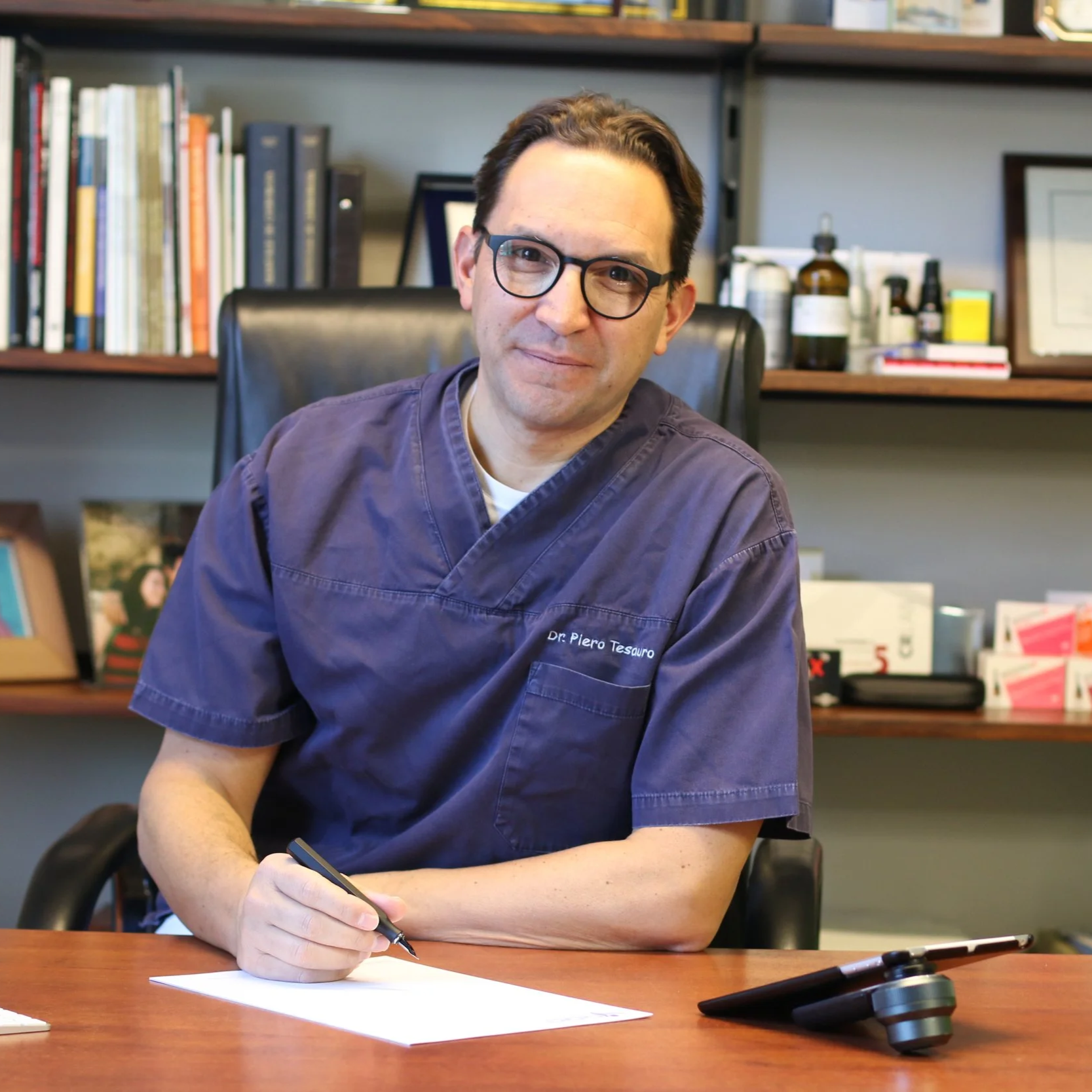 A male doctor wearing glasses and a dark purple scrub top with a name tag reading Dr. Piero Tesauro, sitting at a wooden desk and writing on a piece of paper with a pen. In the background, there are bookshelves with books and medical supplies.