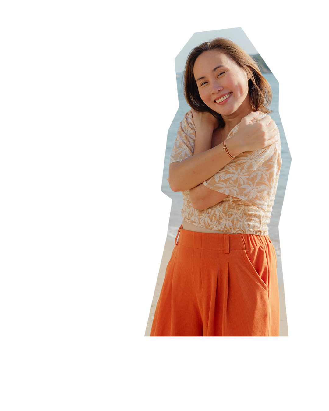 Smiling woman with shoulder-length brown hair standing outdoors near water, hugging herself with crossed arms, wearing an orange skirt and light beige patterned top.