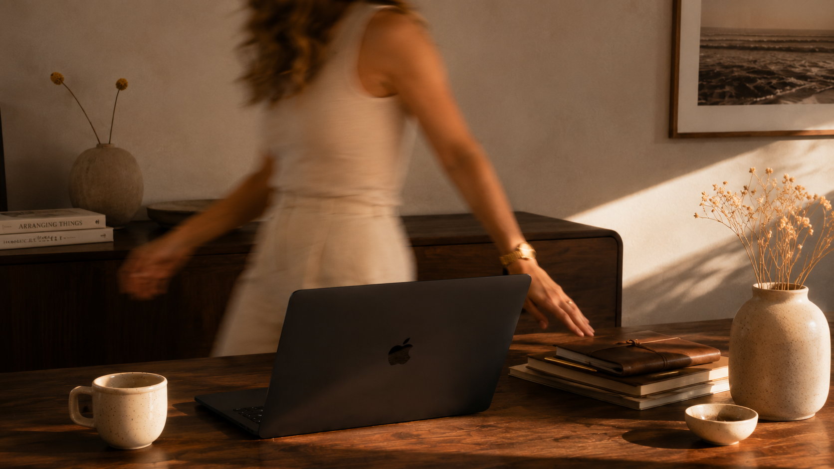 Woman in sleeveless top and skirt walking past a desk with a laptop, mug, and stack of books, in a warmly lit room with decorative vases and framed wall art.